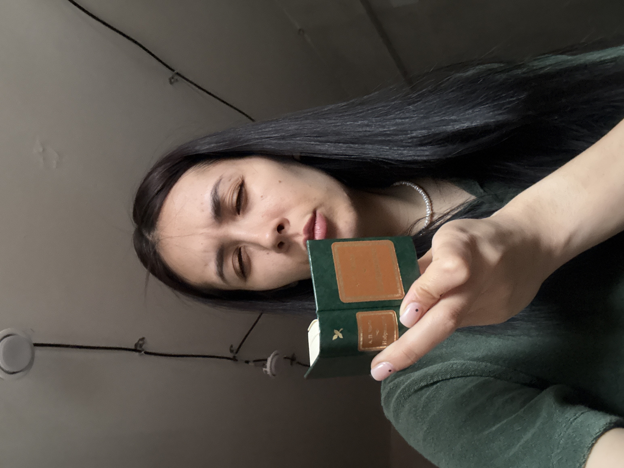 A woman with dark hair and olive skin holds a small, green book with a brown rectangle on its cover, possibly in an indoor setting. The woman is wearing a green shirt, and a pearl necklace is visible on her neck. The photograph appears to have been taken at an unusual angle, perhaps from a lower vantage point looking up. The surroundings are simple, with white walls, visible electrical wiring, and an overhead light fixture, suggesting a residential or office space. Based on the title in Russian, "A.P. Избранное," which means "A.P. Selections", the book could be related to the works of Anton Pavlovich Chekhov. There is no indication that the photograph was taken in Votkinsk, Russia.