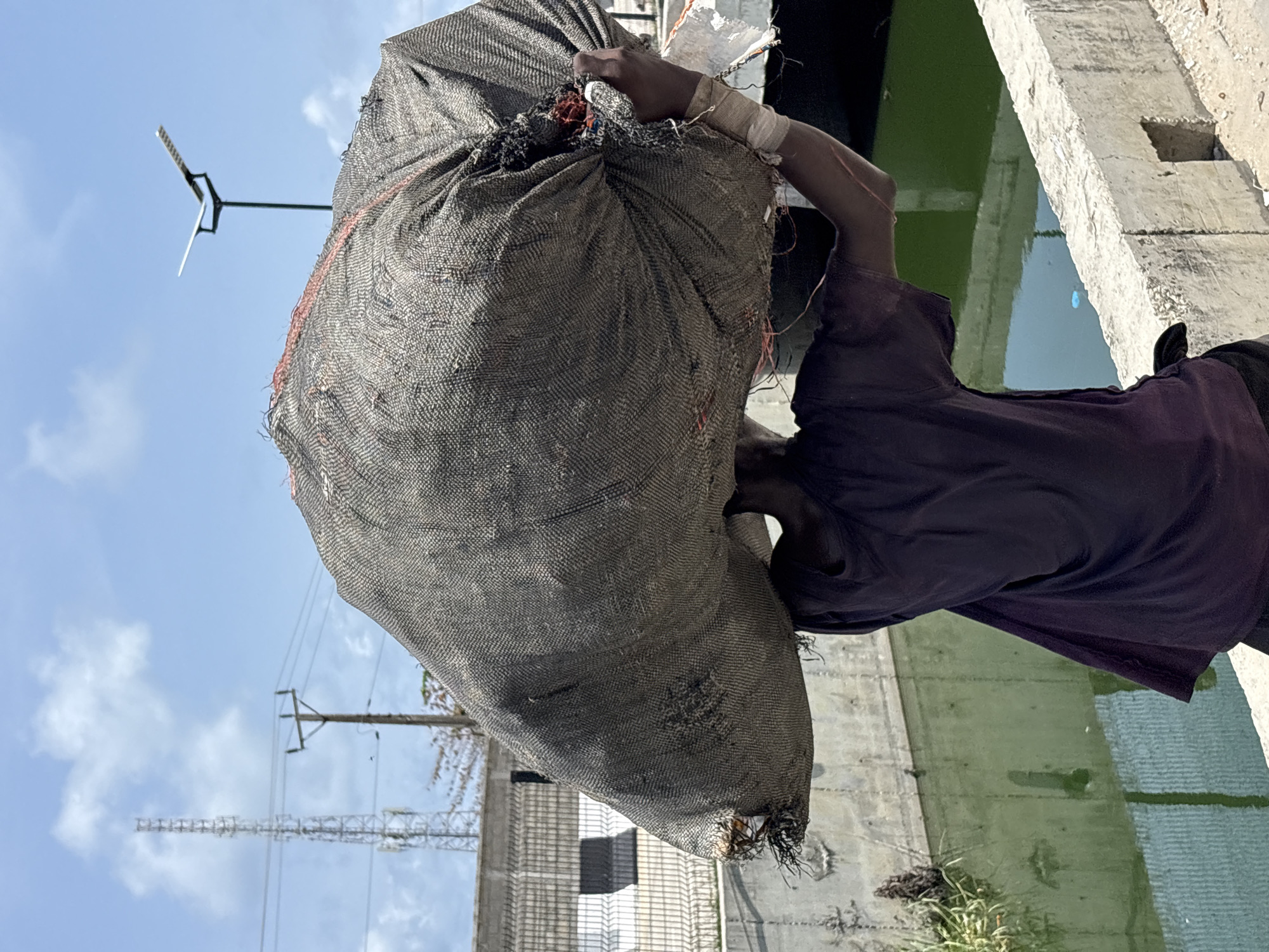 A person, likely an adult male with dark skin, is depicted carrying a very large, heavy-looking, dark grey, coarsely woven bag over their right shoulder. The bag, possibly made of burlap or a similar durable fabric, dominates the foreground and partially obscures the person's head. The person wears a dark short-sleeved shirt, and a white bandage or wrap is visible around their right wrist.
The scene is outdoors under a bright blue sky with scattered white clouds. In the background, electricity poles and power lines stretch across the horizon, with distant buildings and what appears to be construction scaffolding also visible. To the person's right, a concrete structure runs alongside a body of still water, which has a distinct green tint, suggesting algae or industrial conditions. The immediate ground visible near the water is rough and includes some sparse grass. An unusual, small, three-pronged metal object, possibly an antenna or part of a small weather vane, is visible against the sky near the top of the bag.
The activity clearly shows the person engaged in manual labor, transporting a substantial load. The image is oriented such that the person appears horizontal, implying a rotation from the original capture. No visible text is present in the image. The setting suggests an industrial or working area, possibly in Igboefon, Nigeria.
