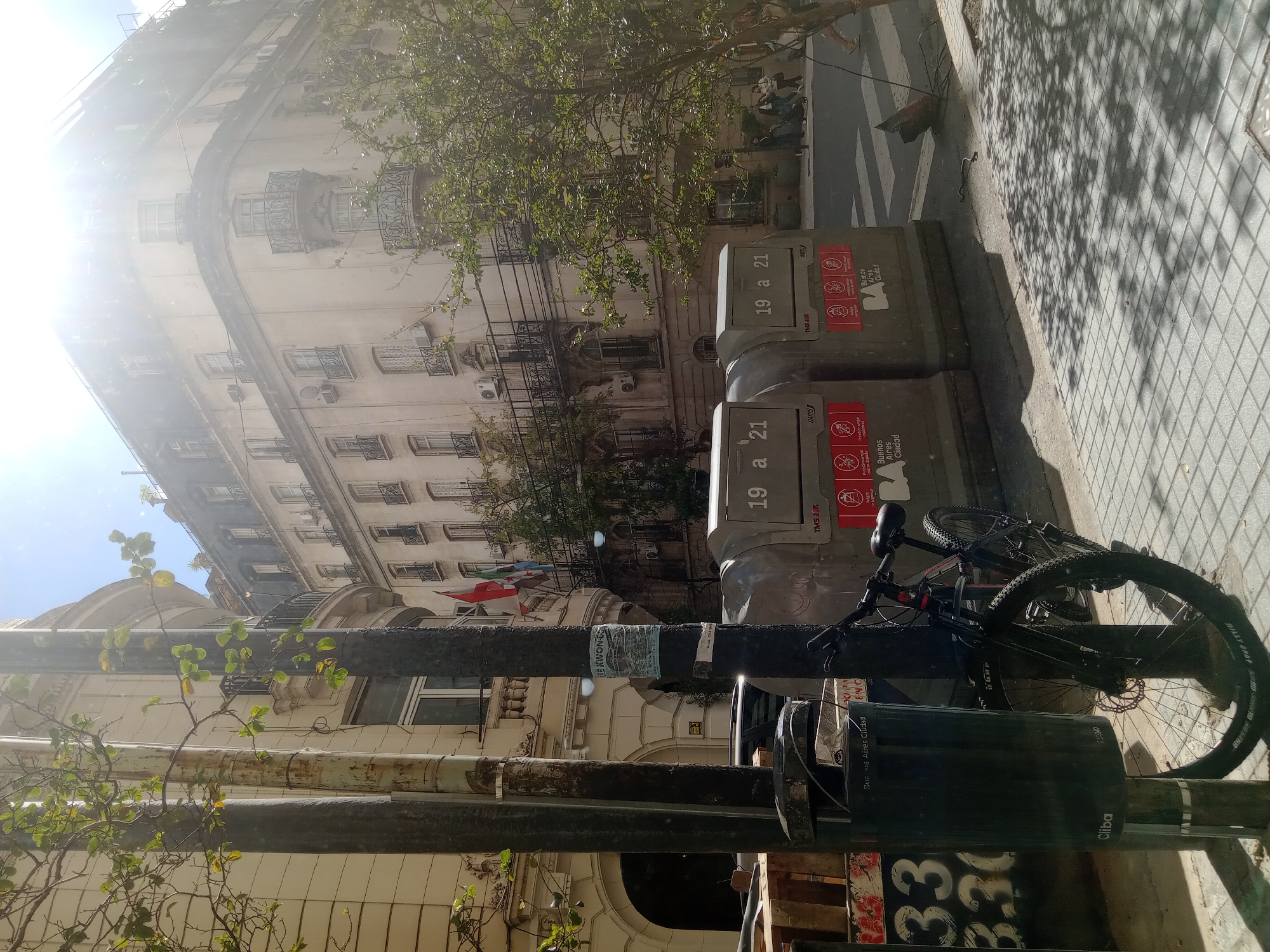 The image captures a street scene in Buenos Aires, Argentina. Two grey, public trash receptacles stand on the sidewalk, labeled "19 a 21" and "Buenos Aires Ciudad." A black bicycle is leaning against a metal post near the receptacles. The number "33" is partially visible on the base of the metal post.
In the background, there are buildings, with a flag flying atop the first building. Sidewalk with a checkered pattern is also visible, with shadows cast from above. The sky is bright. There are trees interspersed throughout the image.