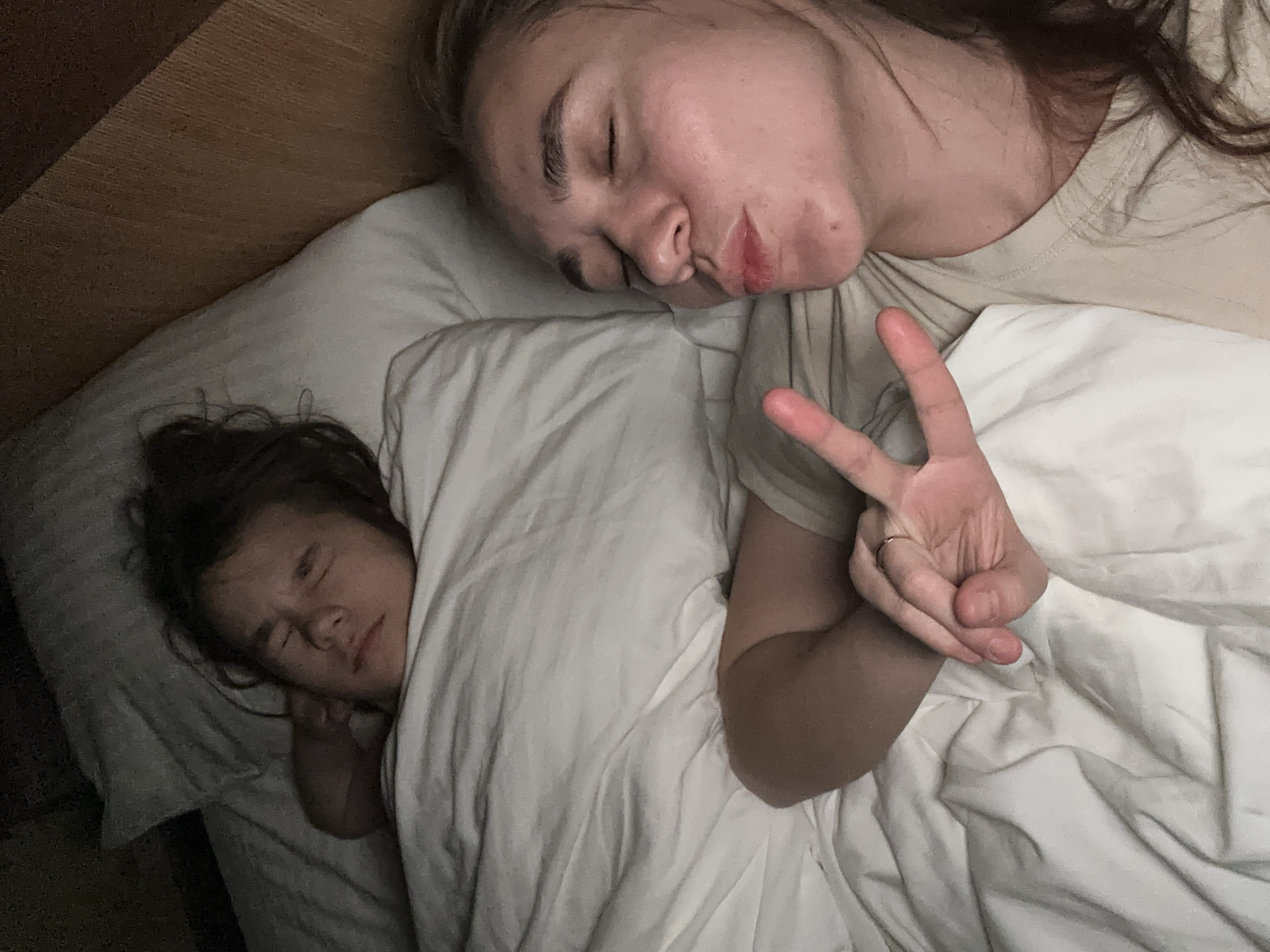 A low-angle, indoor shot shows two individuals lying in a bed. One individual, situated at the top of the frame, appears to be a young adult with closed eyes, light skin, and brownish hair. This individual is making a "peace" sign with their hand. The other individual, lying beneath a white blanket on the left side of the frame, seems to be a child, also with closed eyes and covered partially by the blanket. A small portion of a wooden headboard is visible in the top left corner. The bed is covered in white bedding. No location is identified or implied.