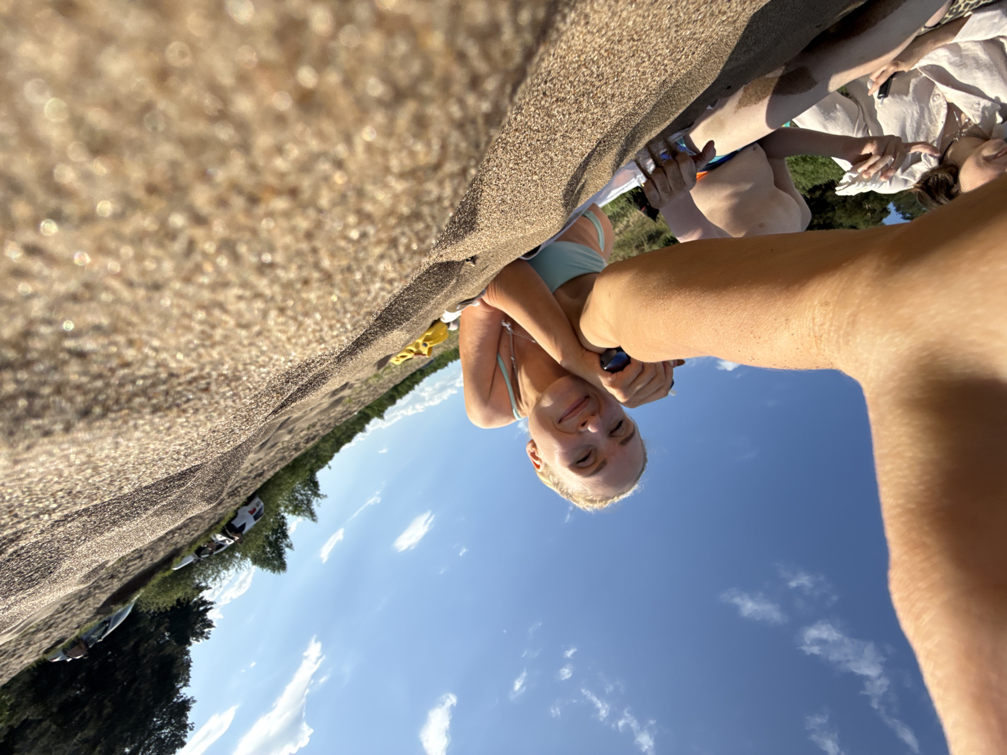 The image is an upside-down selfie taken at a beach, likely in Chaikovsky, Russia, given the context. The foreground is dominated by the edge of a sandy embankment. A young woman with blonde hair is centered in the frame, looking upwards with a slight smile. She wears a light blue bikini top and holds a dark object, possibly a phone, in her hand. Her other arm is positioned across her chest.
Behind the woman, the sky is visible, a clear blue with scattered white clouds. In the far background, green trees line the horizon, suggesting a park or wooded area adjacent to the beach. To the left edge, glimpses of parked cars are visible among the trees. To the upper right, other people appear in partial view, lounging or interacting with each other, one person appears to be wearing a white garment.
