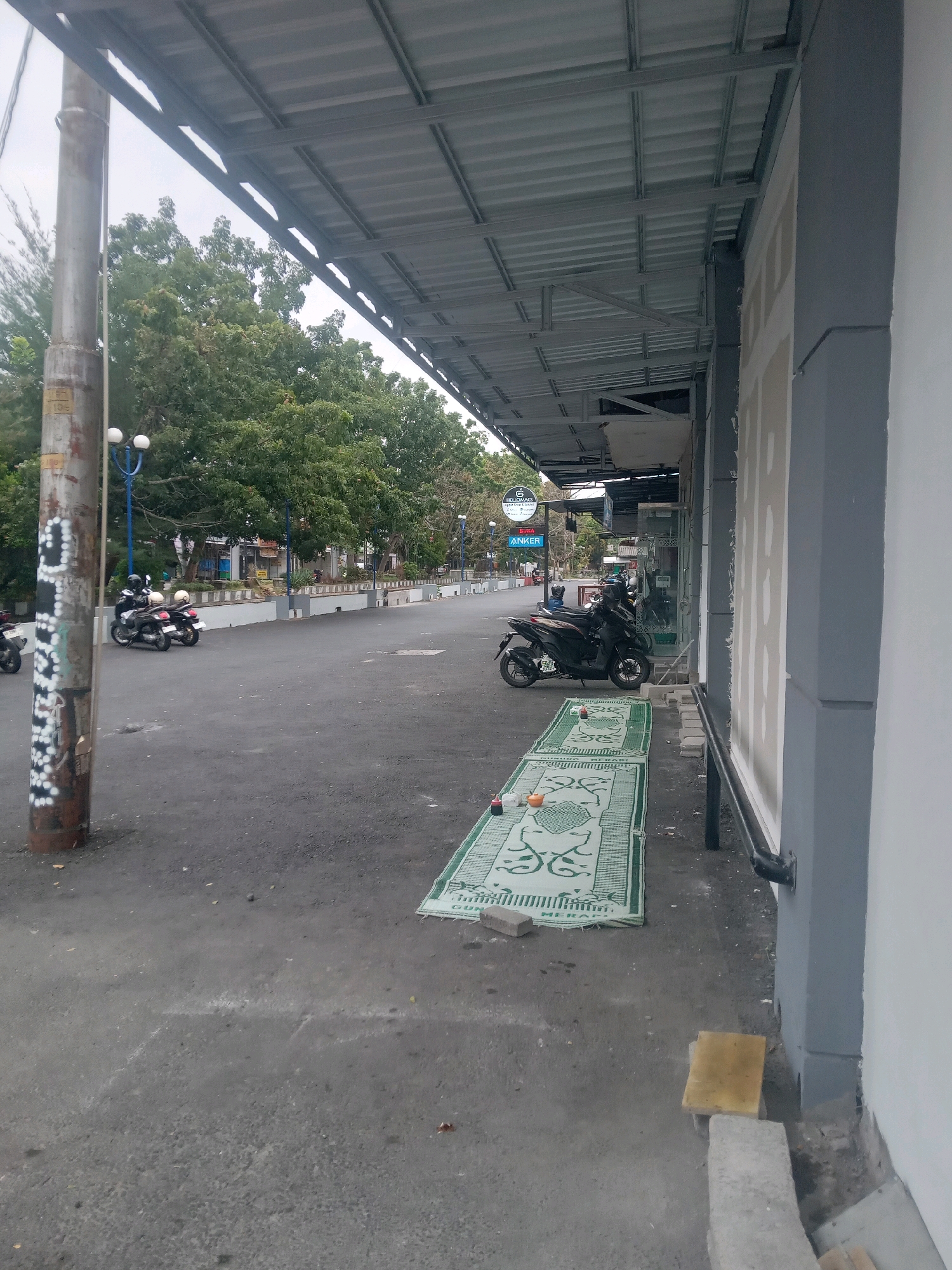 An exterior street-level view in Depok, Indonesia, shows a covered walkway extending along the right side of the image, with a gray, modern building facade to its right. In front of the building, two green prayer mats with intricate patterns are laid out on the asphalt, one with a bottle and two small containers on it. To the left of the walkway, a weathered metal pole stands prominently, adorned with white graffiti and a label. Beyond the pole, a street with a curb separates the sidewalk area from trees and more distant buildings. Parked on the street are several motorcycles, some with riders. Overhead, the corrugated metal roof of the walkway is supported by a metal frame. The sky is overcast.