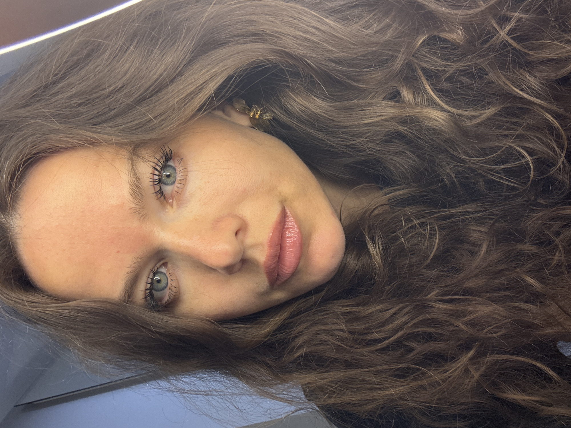A close-up portrait of a woman with long, wavy brown hair and light blue eyes. She has fair skin, light makeup, and is wearing a black top. She has a small, ornate gold earring in her right ear. The background is out of focus, with a light blue wall on the left and a bright light source on the right. The image appears to have been taken indoors, possibly in a residential setting in the city of Khimki, Russia. There is redness on her forehead and cheeks, suggesting she may have a sunburn or be experiencing a flush.