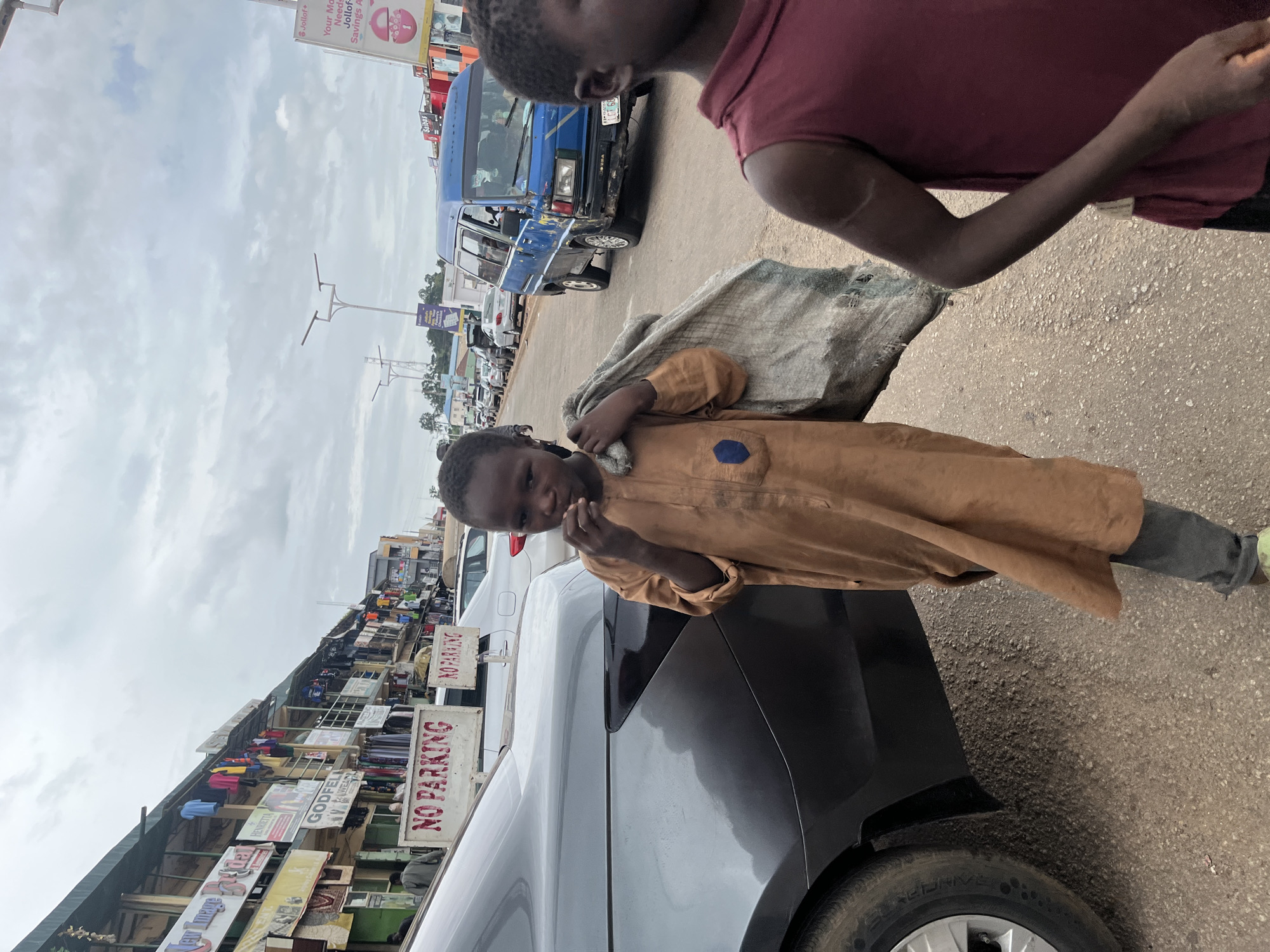 In the city of Jos, Nigeria, an overcast sky looms above a bustling street scene. A young person, dressed in a tan tunic and dark pants, walks forward, carrying a large, grey bundle over their left shoulder. Their right hand is raised to their mouth, and they appear to be eating. Beside them, another young person in a maroon tank top stands partially in the foreground. Cars line the street, including a blue van and a dark grey sedan. Shops with awnings and displays of clothing are visible on the left side of the street, with prominent "NO PARKING" signs. The ground is paved with asphalt and gravel.