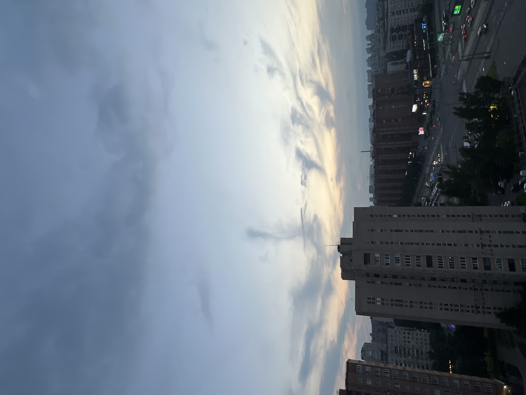 A medium shot captures an urban landscape at dusk, looking out over a cityscape dominated by tall, residential buildings. The sky is a gradient of pale blue and soft grey, with dramatic, wispy clouds that catch the fading light, appearing almost sculptural. Three distinct, thin, dark lines, possibly power lines, traverse the sky diagonally from the upper left to the lower right. In the foreground, a tall, multi-story apartment building with a beige facade and numerous windows stands prominently on the left. To its right, another, darker, and taller apartment building with a similar window pattern is visible. Further in the distance, rows of brick apartment buildings stretch across the horizon, some illuminated by interior lights. A wide street with multiple lanes of traffic is visible in the mid-ground, with cars moving and traffic lights indicating activity. Trees with dark foliage line parts of the street and cluster around the buildings. The overall impression is of a quiet evening in a dense urban environment, with the sky providing a striking natural element against the man-made structures.