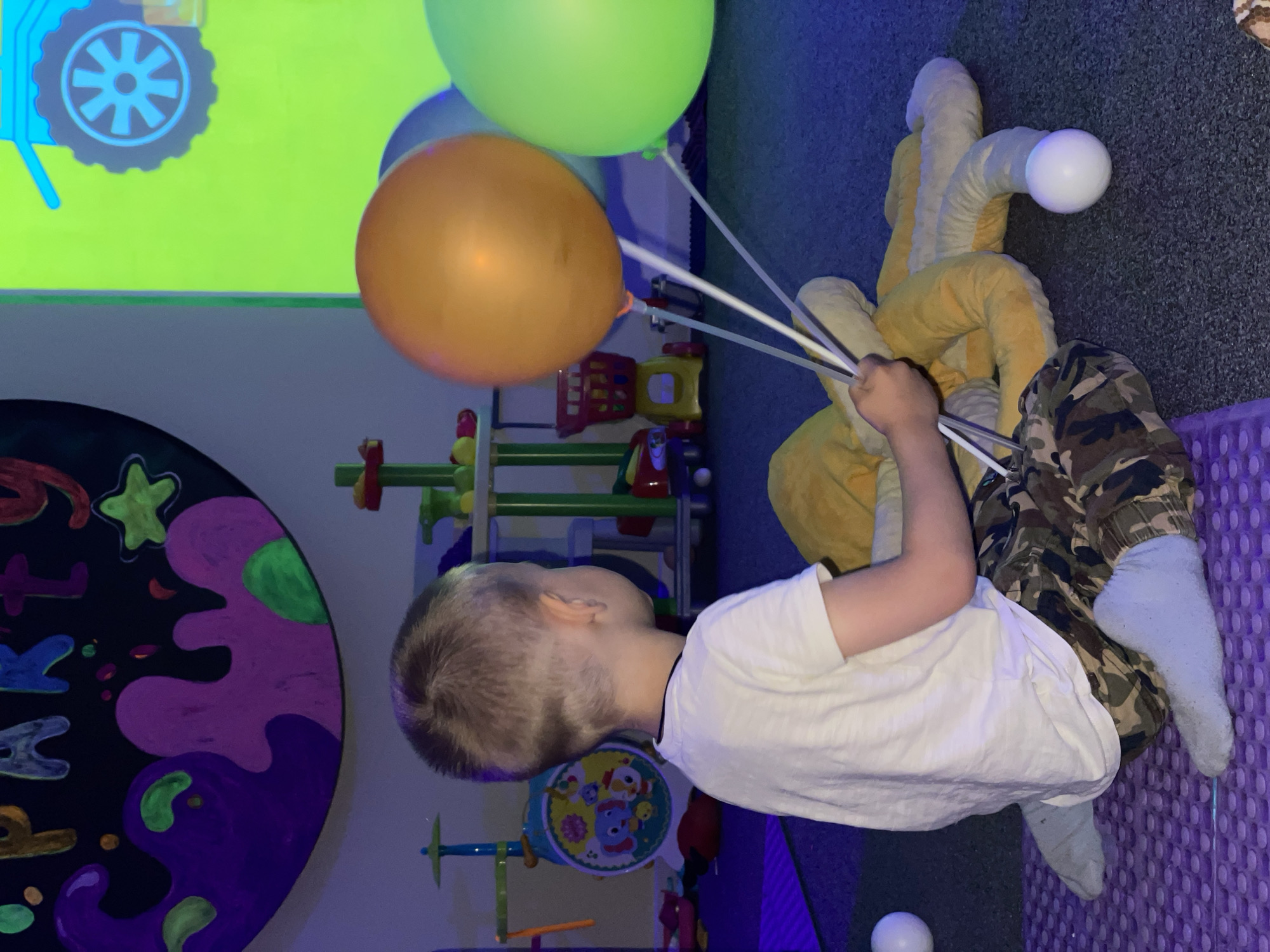 A child in a white t-shirt and camouflage pants holds several balloons in an indoor play area. The balloons are green and orange. The child is sitting on a purple padded surface with a yellow stuffed toy behind them. The play area features a black wall decoration with colorful shapes, a toy shelf, and a toy drum. The room is lit with blue light. The location is Unknown, Unknown.