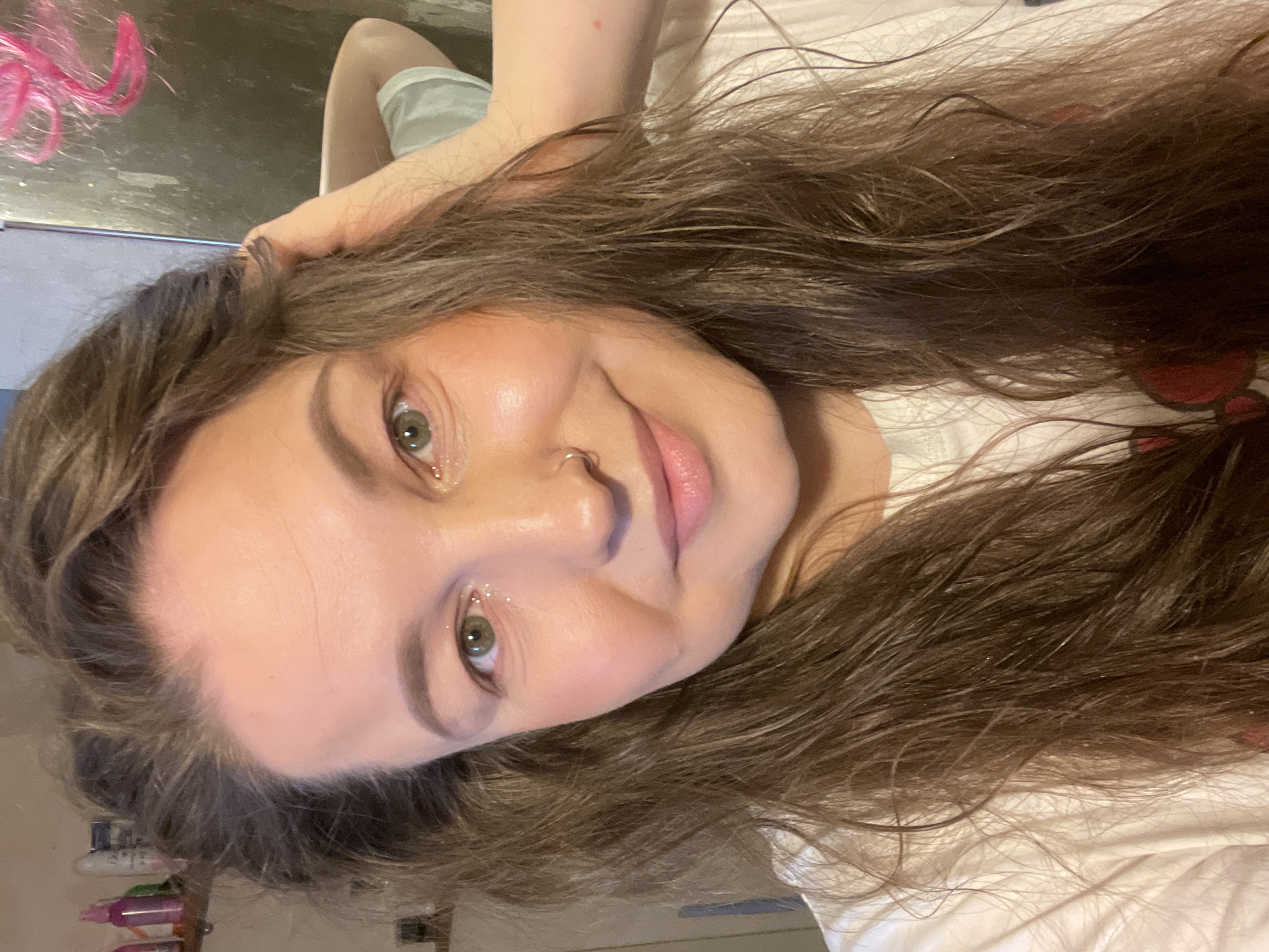 A woman with long, wavy brown hair and green eyes is pictured in a close-up, selfie-style shot. She is wearing a white t-shirt. She has a nose ring and makeup, including glitter eye shadow and blush. Pink hair extensions are partially visible. Bathroom items are in the lower left corner. The image appears to be indoors. Location information is not provided.