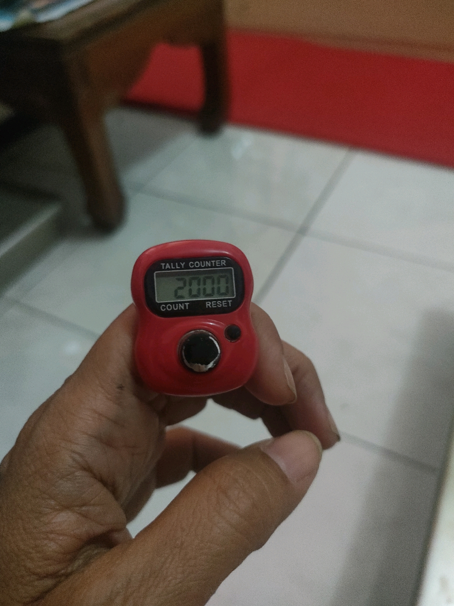 A close-up shot shows a hand holding a red digital tally counter. The counter displays the number "2000". Text on the counter reads "TALLY COUNTER", "COUNT", and "RESET". The background is out of focus, showing a tiled floor and what appears to be furniture.