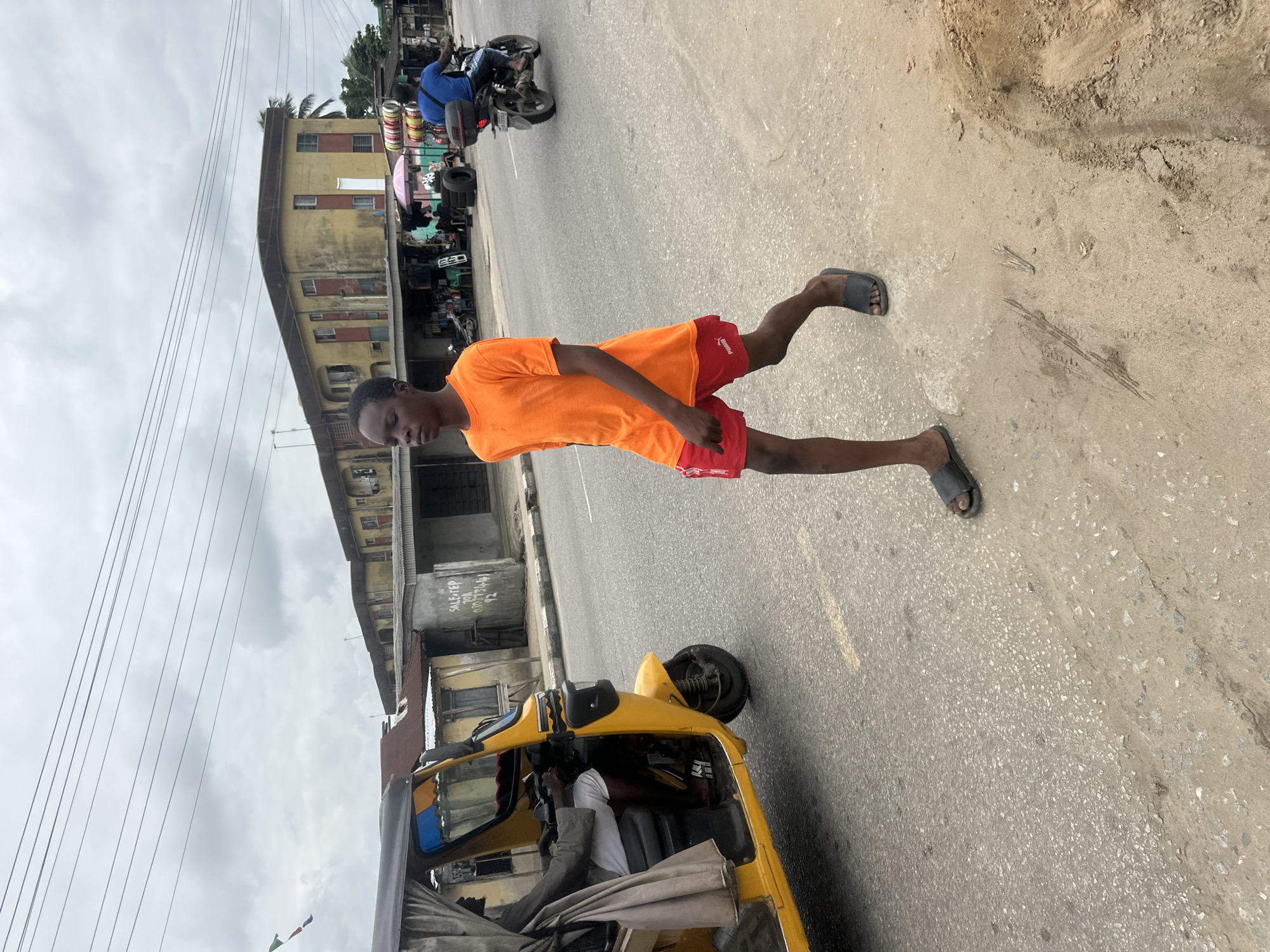 A young boy in an orange t-shirt and red shorts walks on the side of a road in the city of Badagry, Nigeria. He is wearing black sandals. To his left, a yellow auto-rickshaw is stopped. A man is seated in the driver's seat of the auto-rickshaw. Behind the auto-rickshaw, a motorcycle carrying a rider in a blue shirt is moving away from the camera. In the background, there are several buildings, some of which appear to be shops. The sky is overcast.