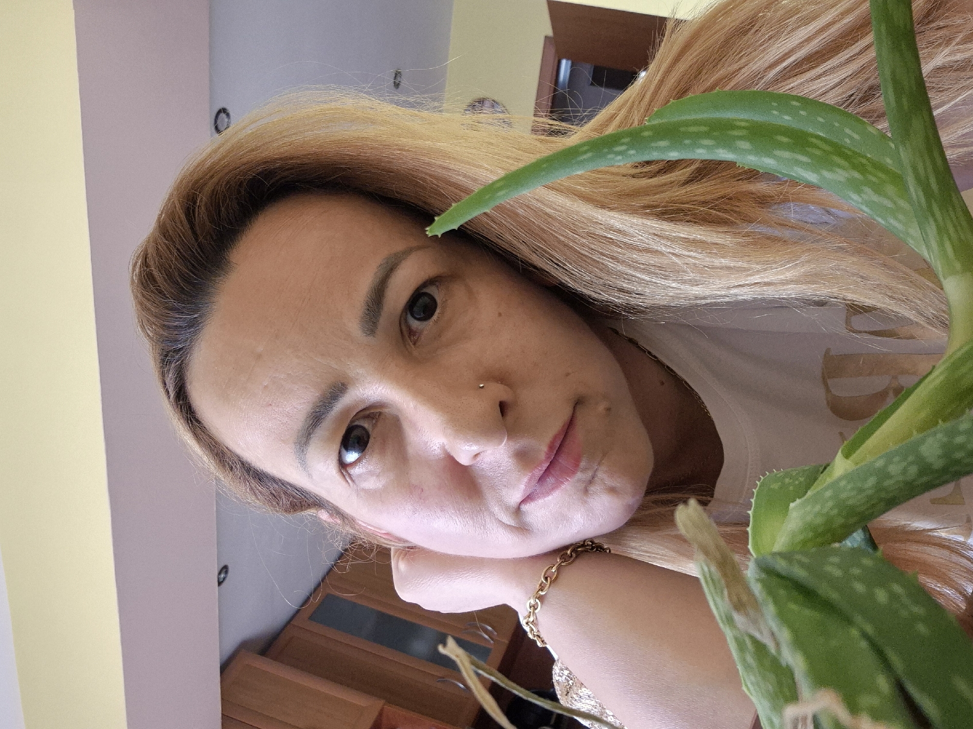 A medium close-up shot captures a woman's face and upper torso, positioned in the foreground and slightly off-center to the right. She has fair skin, light brown hair, and is looking directly at the camera with her left hand resting on her cheek. A gold chain bracelet is visible on her wrist. In front of her, an aloe vera plant with thick, green leaves occupies the lower left and extends diagonally across the frame. The background consists of kitchen cabinetry and a yellow-painted wall, suggesting an indoor setting. The lighting appears to be natural and warm.
