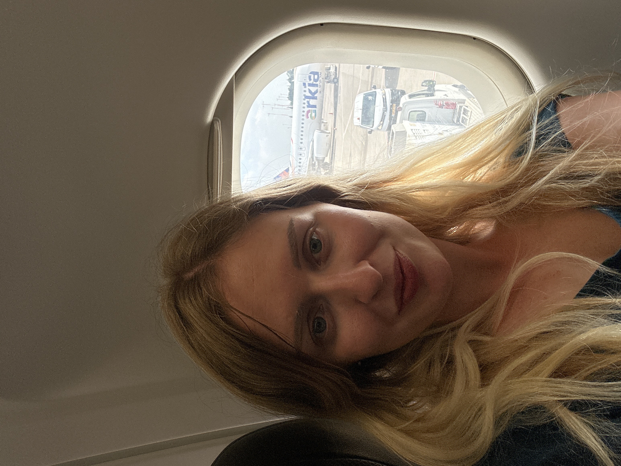 A woman with long blonde hair is seated on an airplane, looking at the camera. She is wearing a dark blue shirt. Through the airplane window, an Arkia airplane is visible on the tarmac, along with service vehicles. The scene appears to be at Ben Gurion Airport in Yehud-Monosson, Israel. The lighting suggests it is daytime.
