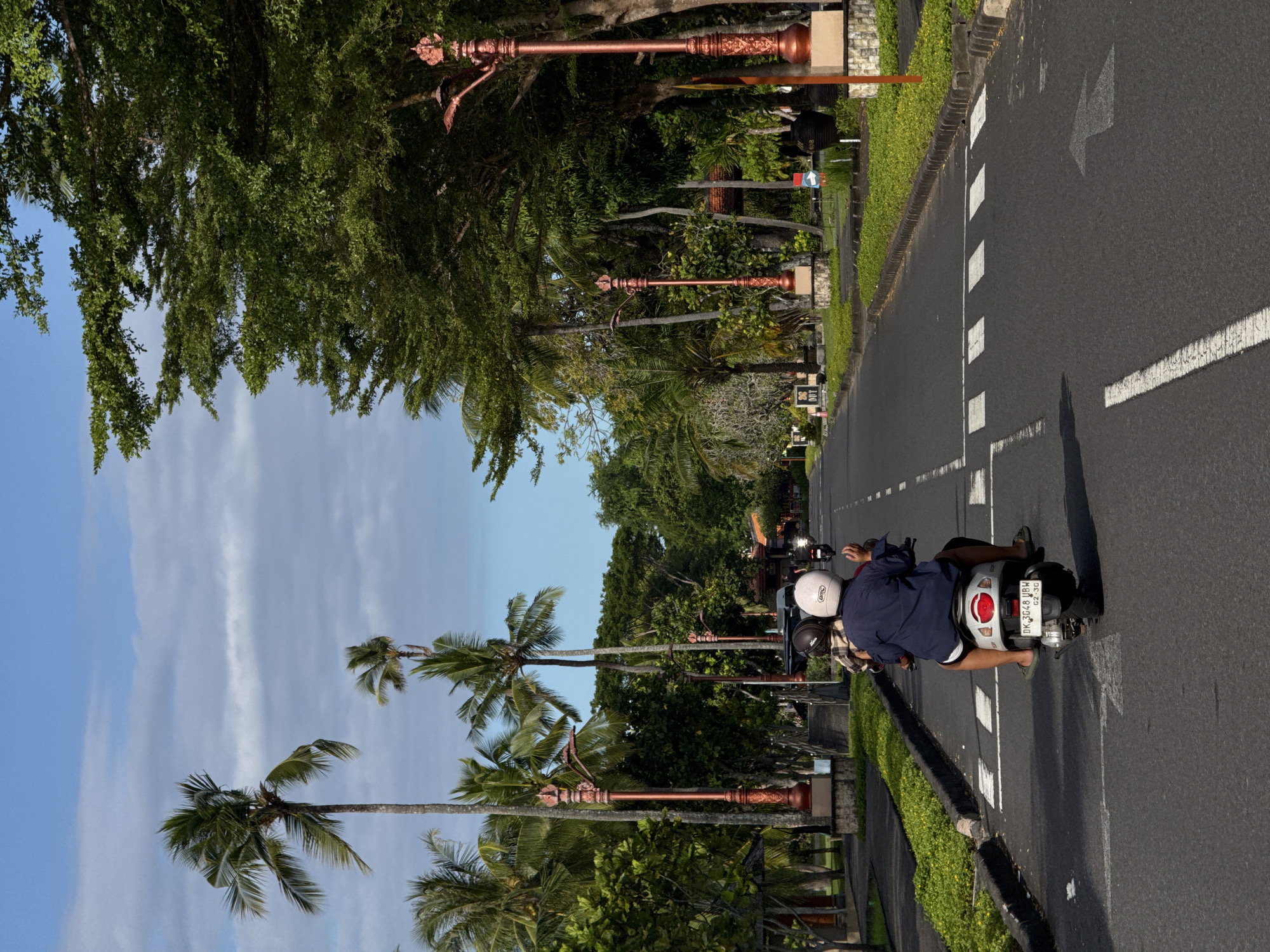 The image shows a street scene in Kuta Selatan, Indonesia. The street is paved and has painted lane markings, including arrows indicating the direction of traffic. Two people are riding a scooter along the road. The scooter has a license plate. The rider is wearing a navy jacket and white helmet, while the passenger is wearing a plaid shirt and black helmet.
The street is lined with lush greenery, including palm trees and other foliage. There are decorative streetlights on both sides of the road. The sky is partly cloudy, with blue patches visible.