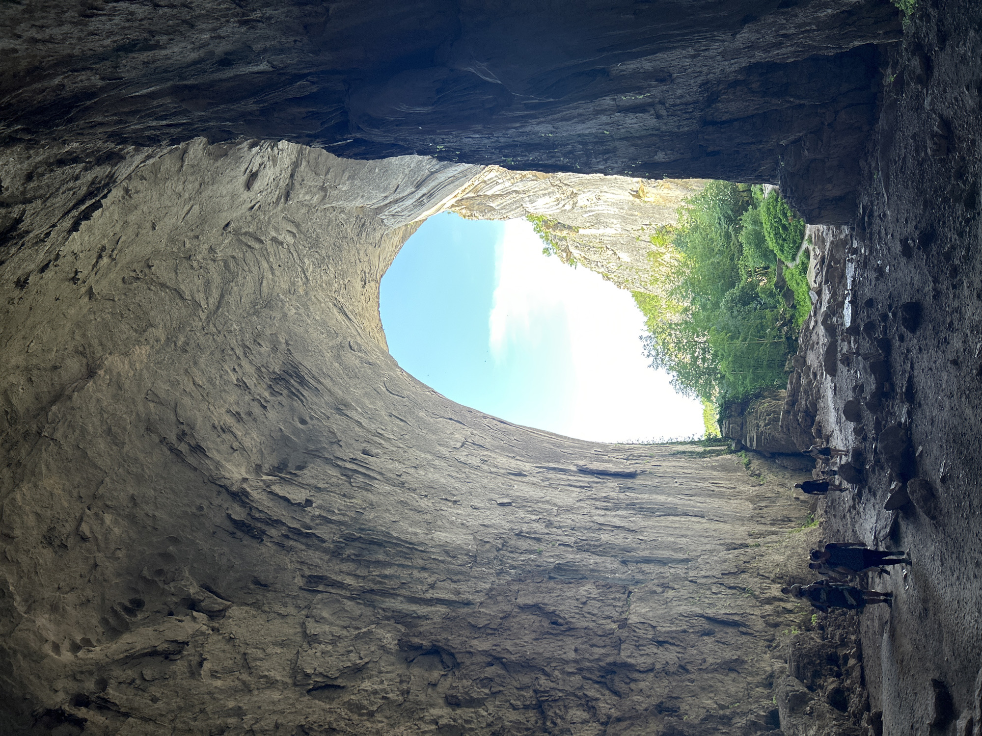 The image shows a view from inside Devetashka cave in Karlukovo, Bulgaria. The angle looks upward through a large opening in the cave ceiling, revealing a blue sky with faint clouds and surrounding green foliage. On the right side of the cave wall, several people are walking along a path. The cave walls are rough and textured, with varying shades of gray and brown. Light enters through the opening, creating brightness at the top and casting shadows within the cave's interior.