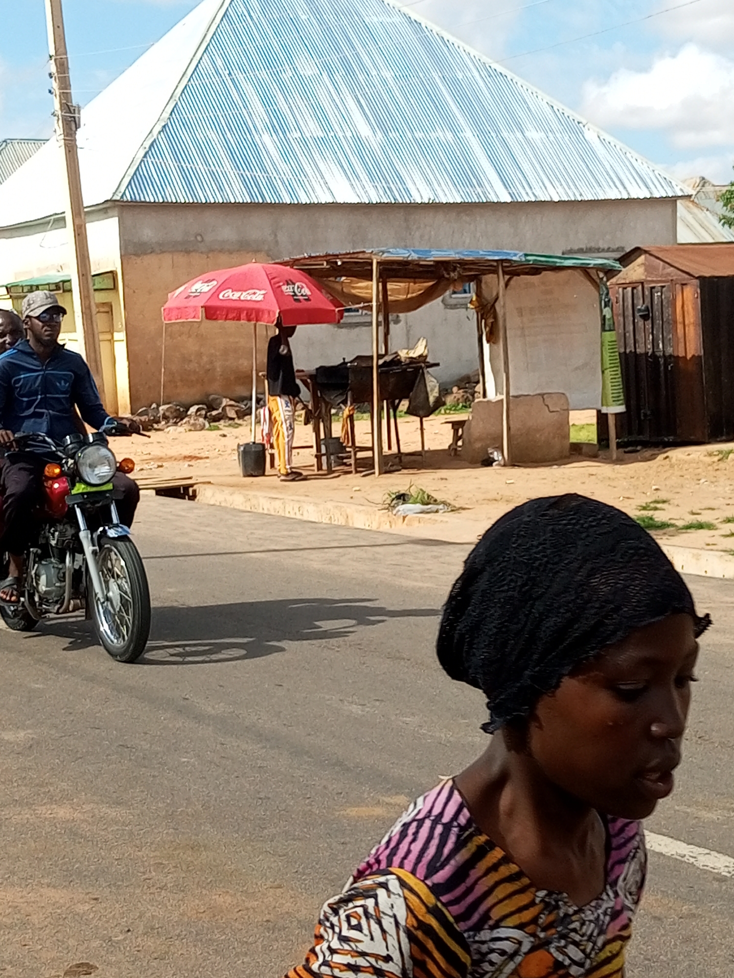 In Tempure, Nigeria, a man wearing a grey cap and dark blue jacket rides a red and silver motorcycle on an asphalt road. He is accompanied by another person partially visible behind him. The motorcycle's shadow stretches across the road. In the foreground, a young woman wearing a black headscarf and a colorful patterned shirt walks on the road. In the background, a red Coca-Cola umbrella shades a small market stall. Behind the stall, buildings with corrugated metal roofs are visible.