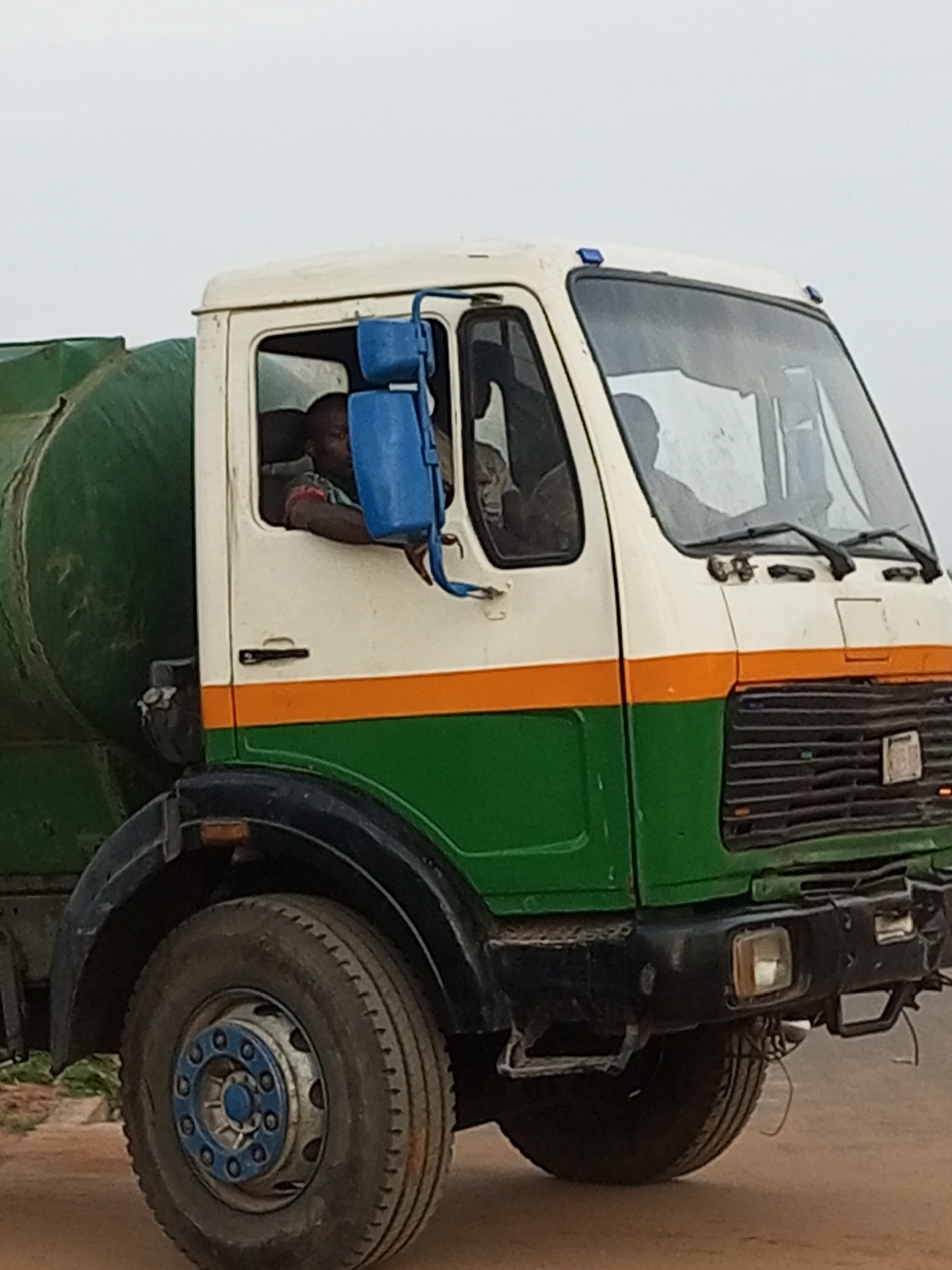 The image shows a large, green and white tank truck with an orange stripe between the green and white sections. A man with dark skin is visible driving the truck, leaning out of the driver's side window. The truck has a large tire with a blue and silver hubcap. The background is a pale, overcast sky. The scene appears to be on a dirt or gravel road. The overall impression is of a functional vehicle in an outdoor setting.