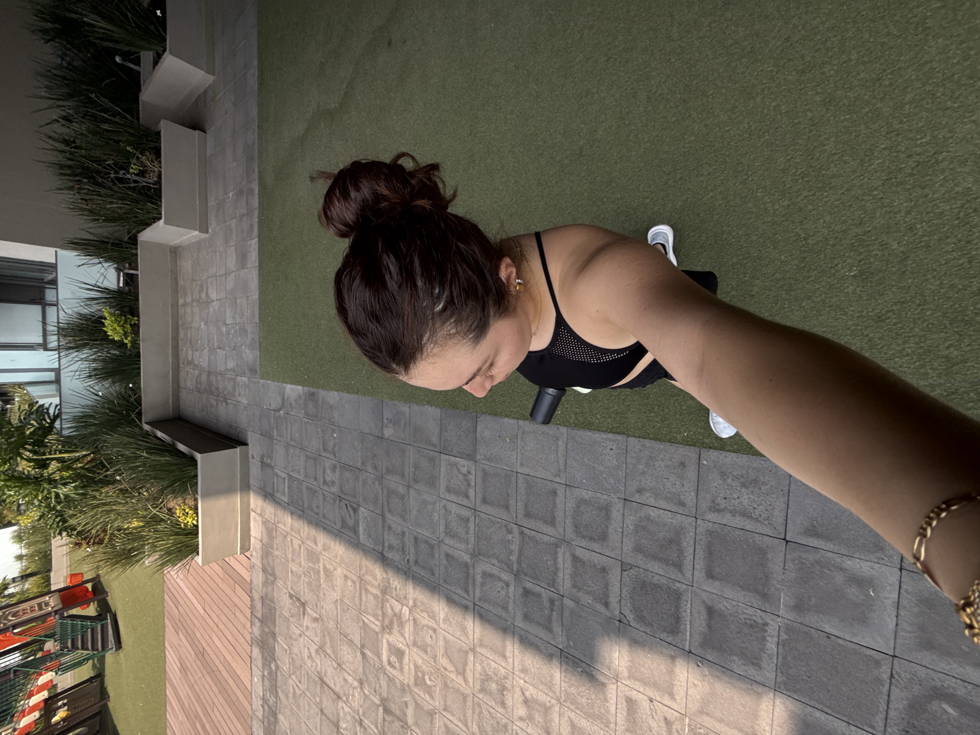 A woman is standing on a paved surface next to a grassy area and a building. She has brown hair in a bun and is wearing a black sports bra. She is holding a black cylindrical object. The background includes a playground area and some plants. The location is likely in Mexico City, Mexico, based on the visible architecture and plants.