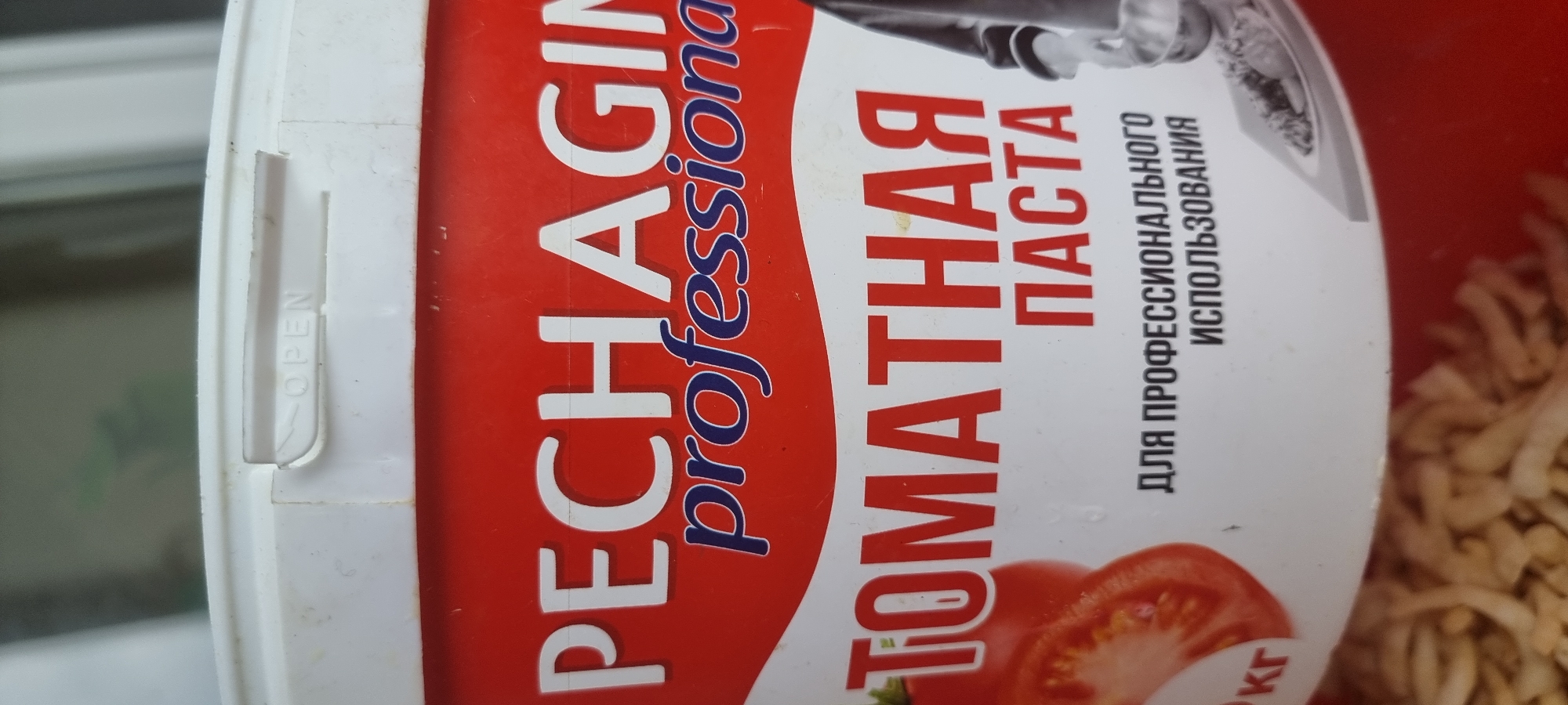 A close-up, slightly angled, vertical shot shows the top portion of a red and white plastic container, likely for food product. The container has text printed on it. The upper rim of the container is white, with the word "OPEN" embossed on a raised section. Below the rim, a red band runs horizontally across the container. Large white text on the red band reads "PECHAGIN" with "professional" in smaller, dark blue script underneath. Further down, larger red text spells out "ТОМАТНАЯ ПАСТА" which translates to "TOMATO PASTE." Below this, in smaller black text, it says "ДЛЯ ПРОФЕССИОНАЛЬНОГО ИСПОЛЬЗОВАНИЯ" which translates to "FOR PROFESSIONAL USE." To the left of this text, a portion of a sliced tomato is visible. To the right, a black and white illustration depicts a hand holding a utensil over a dish of food. The bottom of the image shows the texture of a food product, possibly noodles or shredded vegetables, which appears to be inside or below the container. The scene is lit with artificial light, creating some glare and reflections on the plastic surface. There is no indication of the location being in Aktobe, Kazakhstan.