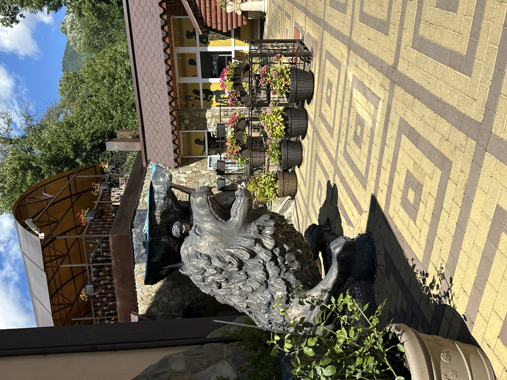 In the city of Krasnaya Polyana, Russia, an outdoor scene unfolds under a blue sky with scattered clouds. The foreground features a large, weathered bronze sculpture of a roaring lion. To the left of the lion sculpture, a large potted plant with green leaves is partially visible. The lion sculpture is situated on a patterned paved area made of yellow and brown rectangular bricks, forming a geometric design of concentric squares.
Behind the lion, a stone wall supports a multi-level structure. The lower level has a brown tiled roof and features a building with yellow walls and large windows adorned with silhouettes of people. The windows are framed by a stone facade. Above this building, a balcony or patio area is visible, supported by metal railings and a yellow awning. Flower pots with red and pink flowers are placed on the railing.
Further in the background, lush green trees and a mountain can be seen. The overall impression is that of a picturesque outdoor seating or dining area, possibly part of a restaurant or cafe, decorated with sculptures and floral arrangements. The lighting suggests it is daytime, with strong sunlight casting shadows from the plant and the lion sculpture onto the paved ground.