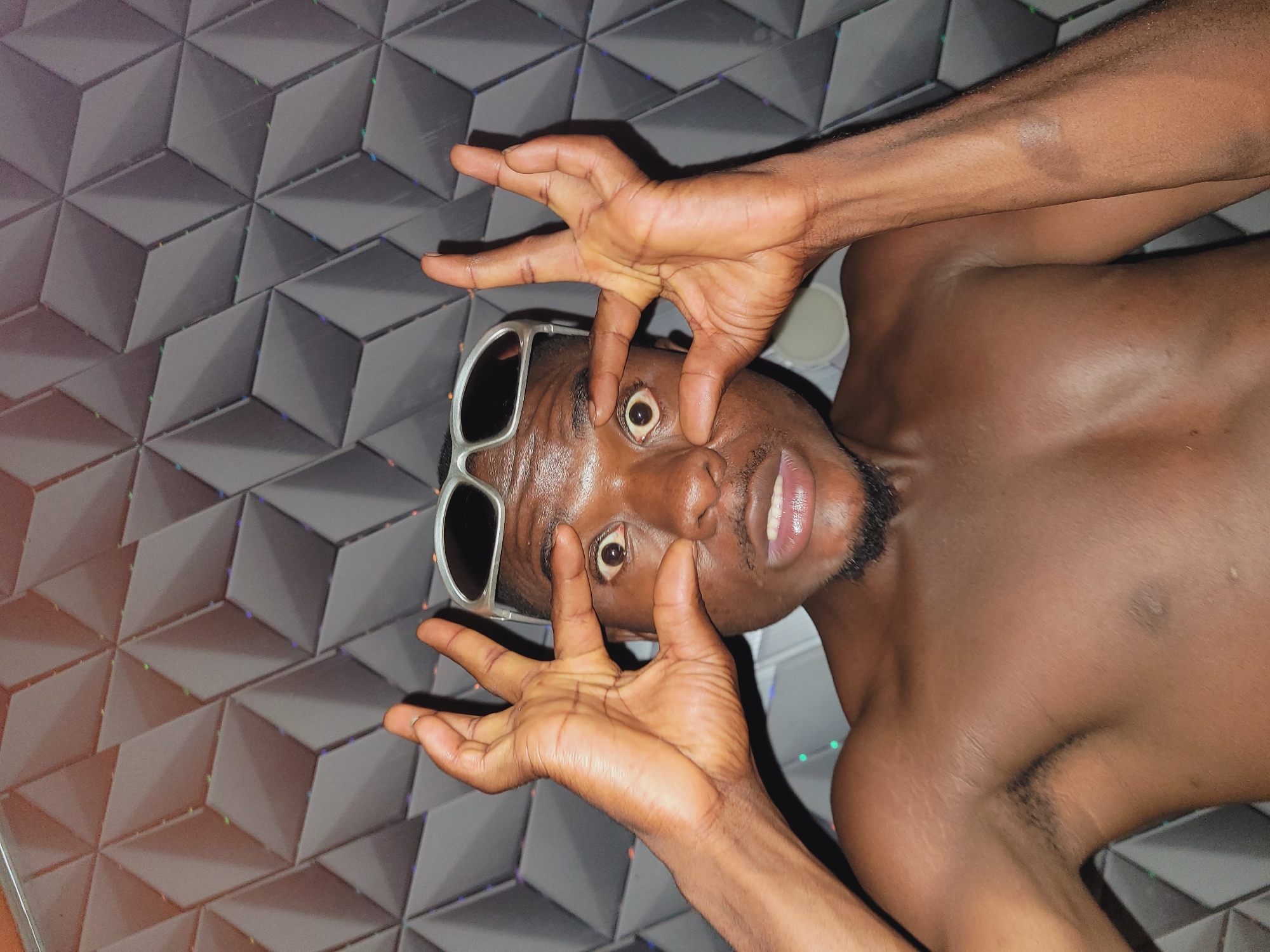 A young Black man with dark skin and a short beard is pictured from the chest up, looking directly at the camera with wide eyes and an open mouth. He is wearing silver-rimmed sunglasses perched on his head. His hands are raised, with his fingers stretched out and touching his eyelids, pulling them wider. The background is a wall with a geometric pattern of raised, dark grey rhombuses. The lighting is dim, with some colorful reflections visible on the wall. The man appears to be shirtless, revealing his chest and shoulders. The image was taken in Bukuru, Nigeria.
