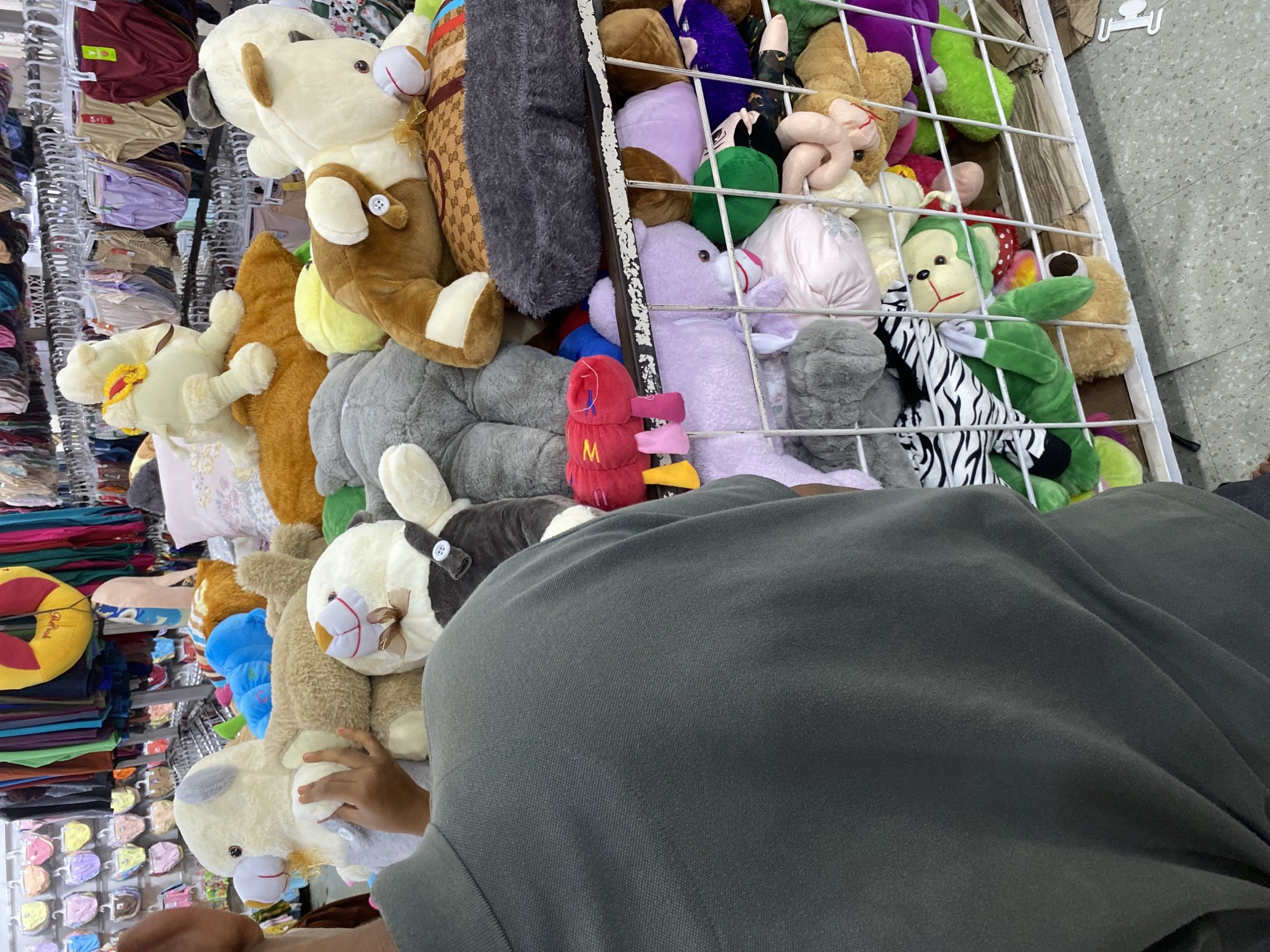 A close-up, over-the-shoulder view of a person wearing a dark green polo shirt is positioned in the foreground, facing a display of numerous stuffed animals. The stuffed animals, primarily teddy bears and other cartoonish creatures, are densely packed within a white metal shelving unit. The stuffed animals vary in color, size, and style, with some featuring ribbons, bows, and buttons. In the background, the retail environment is visible, showing racks of clothing, including undergarments, and colorful fabrics hanging from hangers. The lighting suggests an indoor retail space. This scene likely depicts a merchandise display within a store located in Lamteumen Barat, Indonesia.