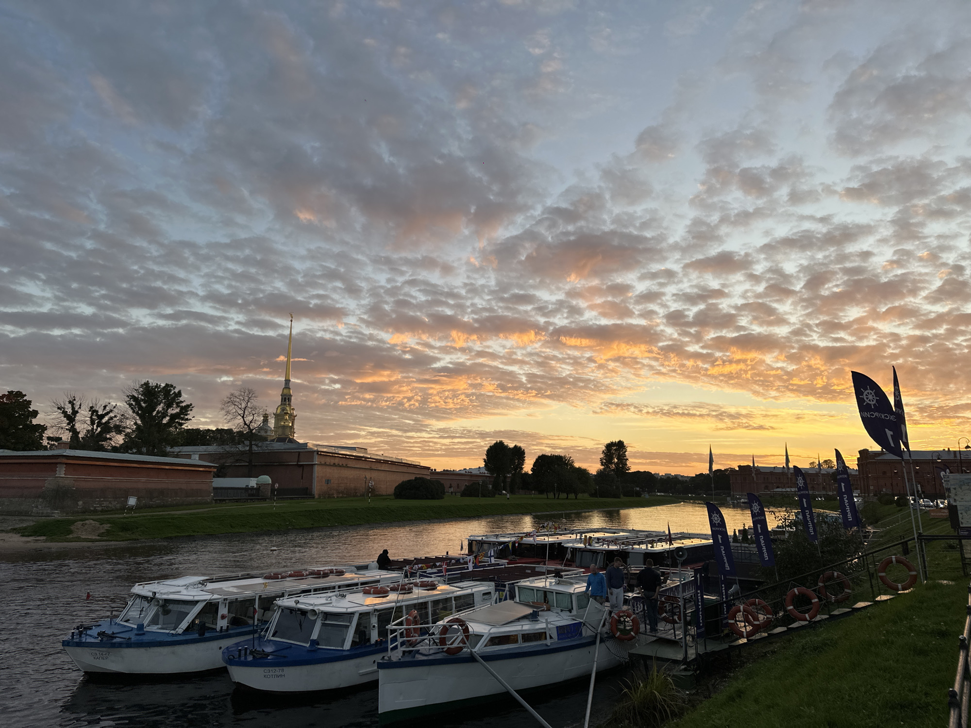 The image depicts a sunset over the Neva River in Saint Petersburg, Russia, with a line of tour boats docked along the embankment. The sky is filled with dramatic clouds, tinged with orange and pink from the setting sun. On the far bank of the river, the Peter and Paul Fortress, a prominent landmark, is visible with its distinctive spire. Several tour boats are moored together, some with people on board, suggesting a tourist excursion. The embankment is lined with grassy areas and a few trees. Dark flags with a nautical wheel emblem are displayed on poles along the dock. The water is calm, reflecting the colors of the sky.