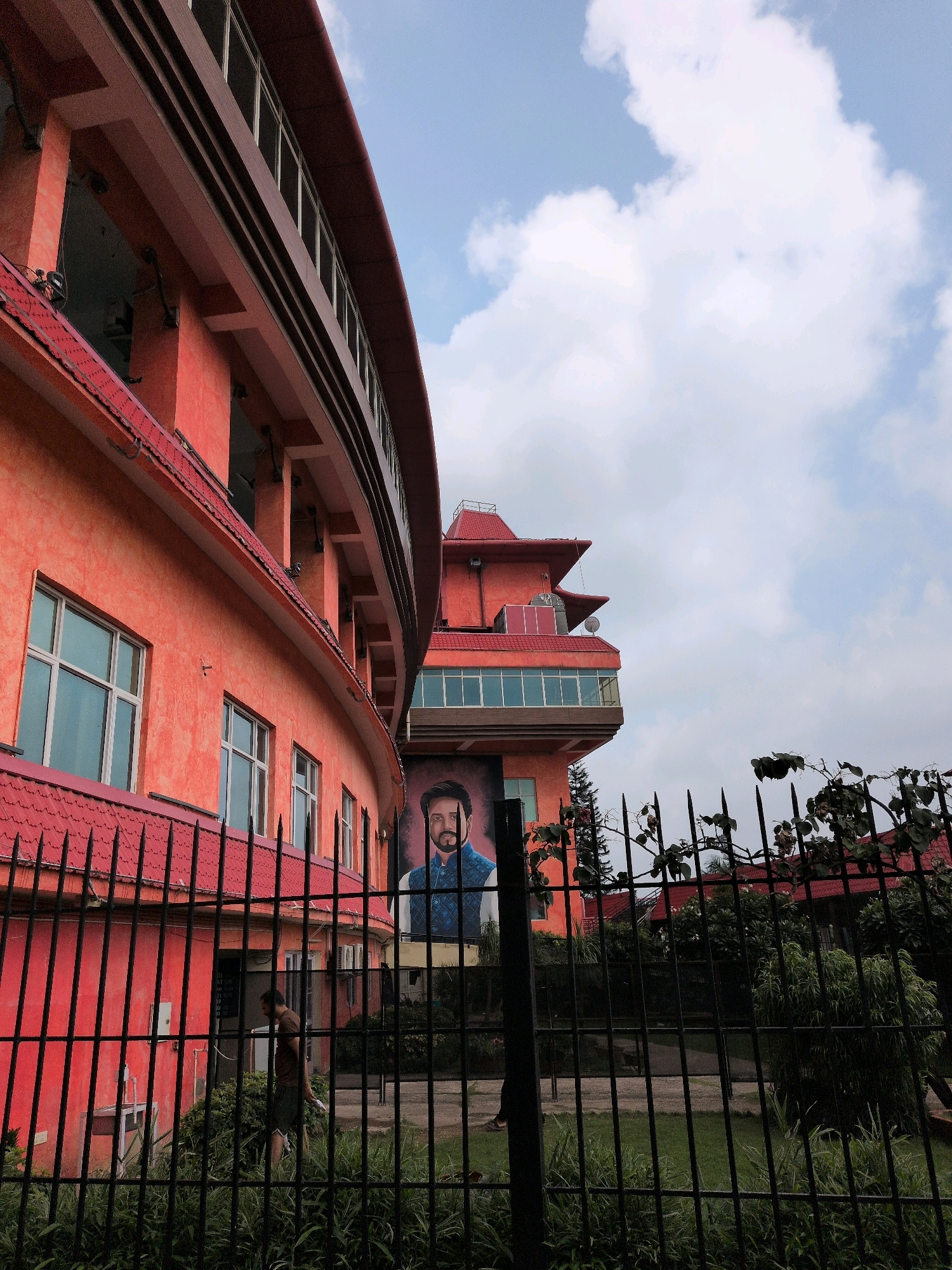 The image captures a frontal view of a large, curved, coral-orange building with red tiled roofs, possibly a hotel or institutional complex, located in Dharamshala, India. The building features multiple stories with arched window sections and decorative awnings. Prominently displayed on the building's facade is a large portrait of a man wearing a traditional blue vest over a white shirt. A black metal fence, topped with pointed spikes, runs across the foreground, partially obscuring a grassy area and some foliage. Behind the fence, a person in a brown shirt and dark shorts is walking on a paved path. The sky above is a mix of light blue with scattered white clouds. The architecture and the man's attire suggest a South Asian context, and the mention of Dharamshala places the scene in the northern Indian state of Himachal Pradesh.