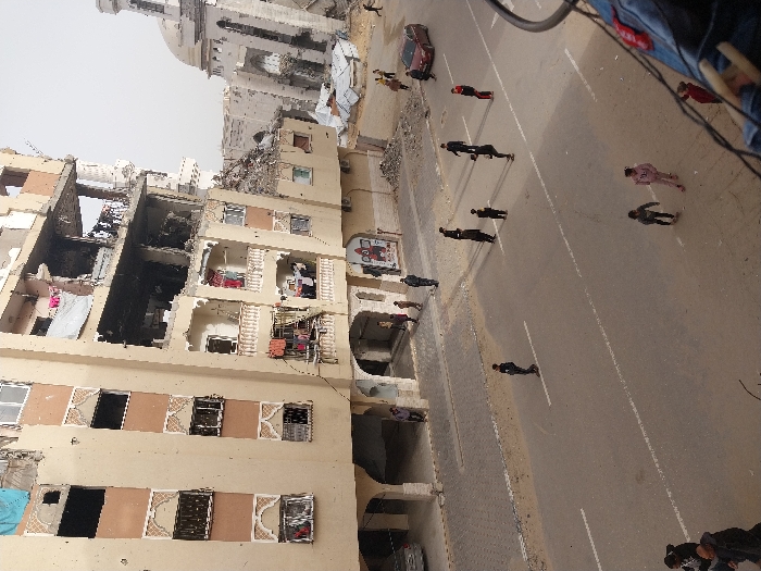 An elevated view captures a street scene in Khan Yunis, Palestinian Territory, characterized by extensive structural damage to multiple urban buildings under an overcast sky.
The prominent structure on the left and central-left is a multi-story, beige-colored residential or commercial building, showing significant damage. Its upper floors display missing windows, exposed interiors, and large sections of collapsed walls, particularly on the upper-right section, revealing blackened areas possibly from fire. Balconies with white balustrades are visible on lower and mid-levels, some draped with laundry. The ground floor features arched entryways and underpasses.
To the right and behind this building stands a heavily damaged mosque-like structure with a light-colored, partially intact dome and a slender, damaged minaret. The area between the beige building and the mosque is strewn with rubble and debris, some covered by white tarpaulins.
A grey asphalt road with faded white road markings (double solid lines in the center, dashed line on the right) bisects the scene. Approximately 15-20 individuals, mostly adults and older children, are scattered across the street, either standing or walking. Smaller groups are visible near the building entrances and further down the road. Two younger individuals in lighter clothing are in the lower foreground.
A red sedan is parked on the right side of the road, facing away from the viewer, and a silver car is partially visible beneath an archway of the beige building on the left. The overall scene depicts a damaged urban environment with ongoing human presence.