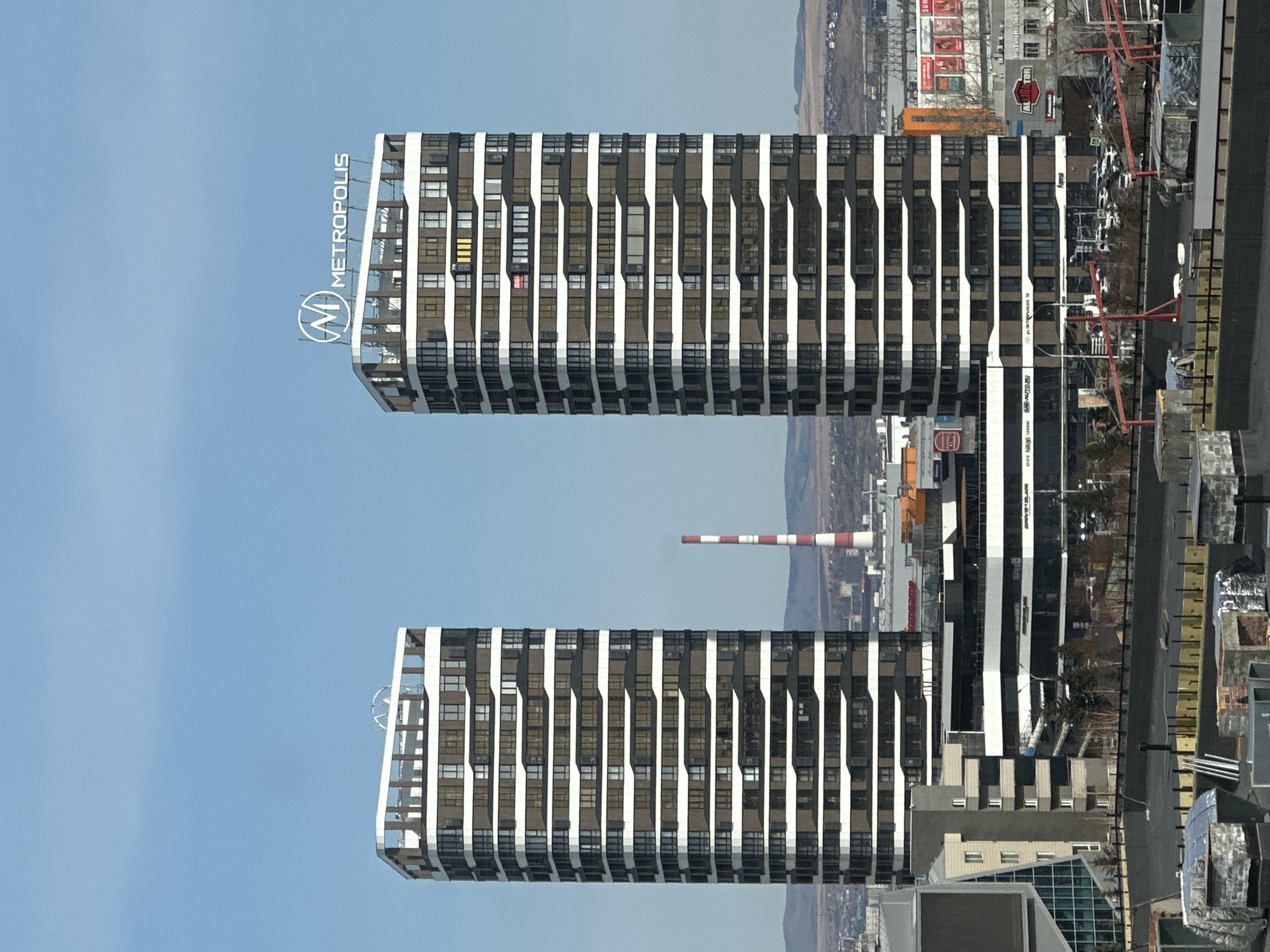 This is an exterior shot of the Metropolis apartment complex located in the city of Krasnoyarsk, Russia. The image shows two towers of the complex, characterized by a repeating pattern of dark-framed windows with white vertical supports. The towers are situated in a built-up urban area, with other buildings and infrastructural elements visible in the background. The sky is clear blue. The "METROPOLIS" logo is visible on top of the top tower.