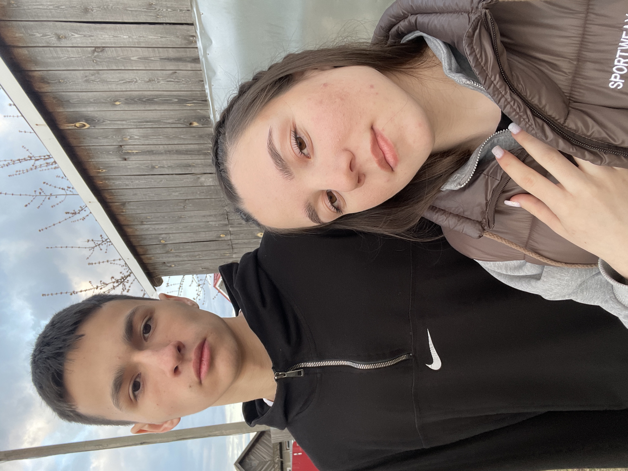 This media file captures two young individuals, a male and a female, in a tilted portrait outdoors in what appears to be Ольховка, Russia. The female, positioned closer to the camera in the upper frame, has dark hair styled with a braid along the hairline and wears a brown puffer vest over a grey hooded top. The word "SPORTWEAR" is visible on her vest, and her nails are painted a light, possibly glittery, color. Her complexion shows some natural redness. The male, in the lower part of the image, has short dark hair and wears a black hooded jacket with a prominent white Nike swoosh logo on the chest. Both individuals maintain neutral to slightly serious expressions while looking towards the camera.
The background features a weathered wooden fence or wall made of horizontal planks. On the left, a white horizontal beam or railing is visible, with bare tree branches silhouetted against an overcast or cloudy sky. A small building with a red roof can be faintly discerned in the lower right background. The scene suggests a daytime setting under subdued natural light due to the cloudy weather. The overall impression is a casual outdoor moment.