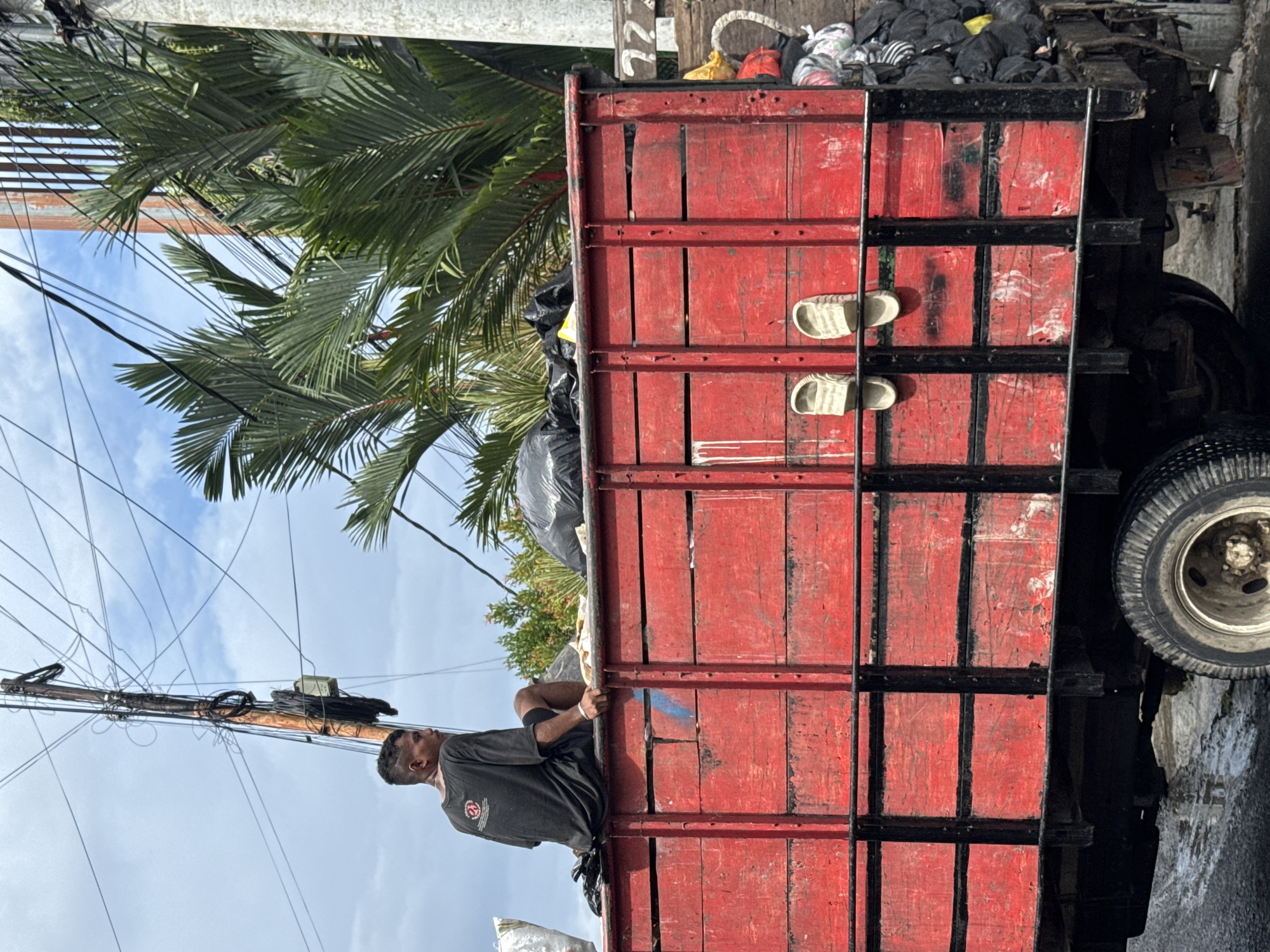 In North Kuta, Indonesia, a utility worker climbs onto the back of a red, black-framed truck laden with black trash bags. The truck, positioned on a wet asphalt road, has two pairs of white sandals tucked into its frame. Overhead, a dense cluster of power lines intersects with a tall palm tree under a cloudy sky.