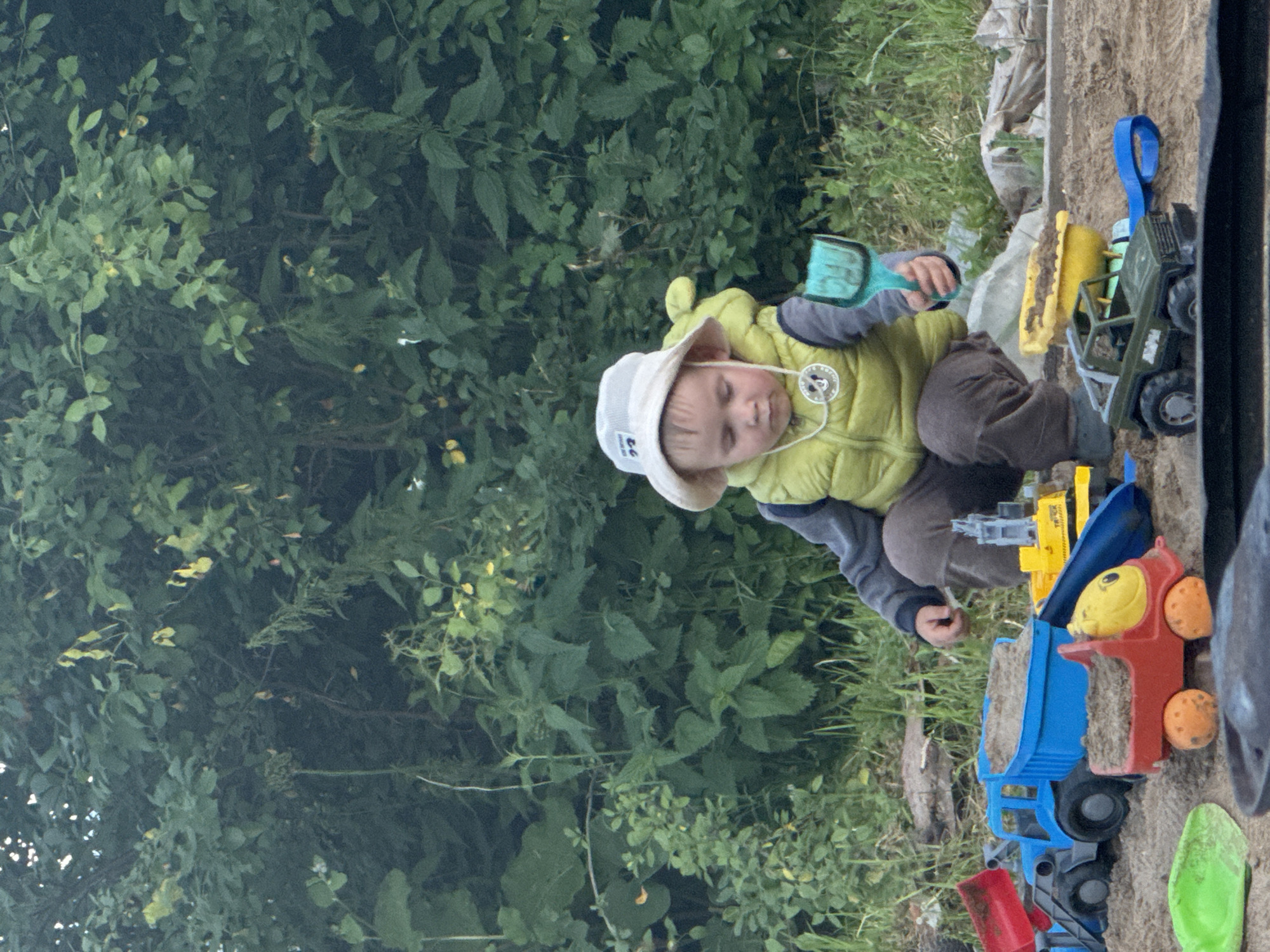 In a natural outdoor setting, possibly a yard or park in the city of Prikamsky, Russia, a young boy is engaged in play. He is wearing a light-colored bucket hat and a bright yellow vest over a grey long-sleeved shirt and brown pants. The boy is crouched down in a sandbox, holding a turquoise toy shovel. Around him are several colorful plastic toy construction vehicles, including a blue dump truck filled with sand, a red toy truck, a yellow toy digger, and a green toy jeep. The sand is spread across the ground, and in the background, there is dense green foliage and trees, suggesting a day spent outdoors.
