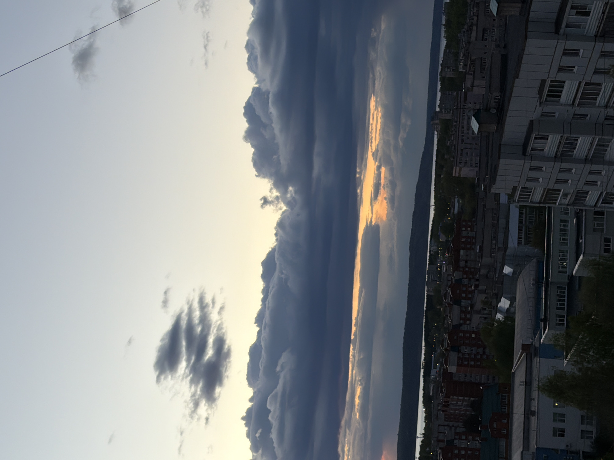 The image shows a cityscape in Chaikovsky, Russia, likely taken from a high vantage point. A body of water (possibly a river or large lake) separates the foreground city from a darker landmass in the background. The sky is dominated by dark, heavy clouds, with a patch of golden light breaking through near the horizon. Buildings are visible in the foreground.
