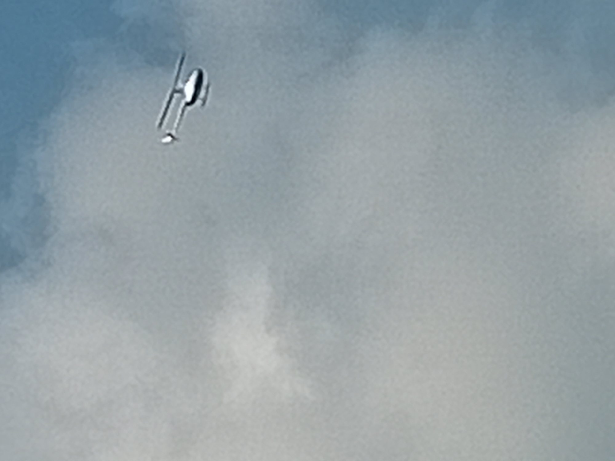 A small, light-colored helicopter flies in the sky. It has a single rotor with two blades, and a tail rotor. The helicopter is silhouetted against a cloudy sky. The clouds are light gray and white, and the sky is a pale blue. The helicopter appears to be in flight over Kuta, Indonesia.
