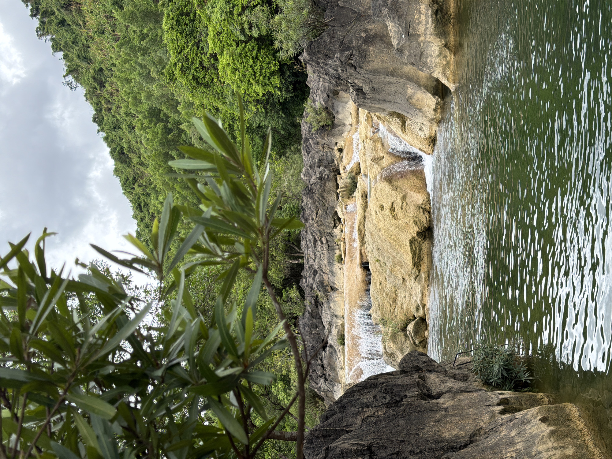 This image captures a natural landscape in Trường Sơn Commune, Vietnam, featuring a tiered waterfall cascading over light-colored rock formations into a tranquil, greenish body of water. The foreground is partially obscured by the dark, leafy branches of a tree. On the left, a large, dark gray boulder juts into the water. The middle ground displays the waterfall, with water flowing over tan and yellowish rocks, creating a frothy white spray. Several smaller streams and pools are visible among the rocks. In the background, a lush, green, tree-covered hillside rises under a cloudy sky. The overall scene is one of natural beauty and serenity, with no people or human-made structures immediately apparent besides the hints of a rough path or structure near the water's edge.