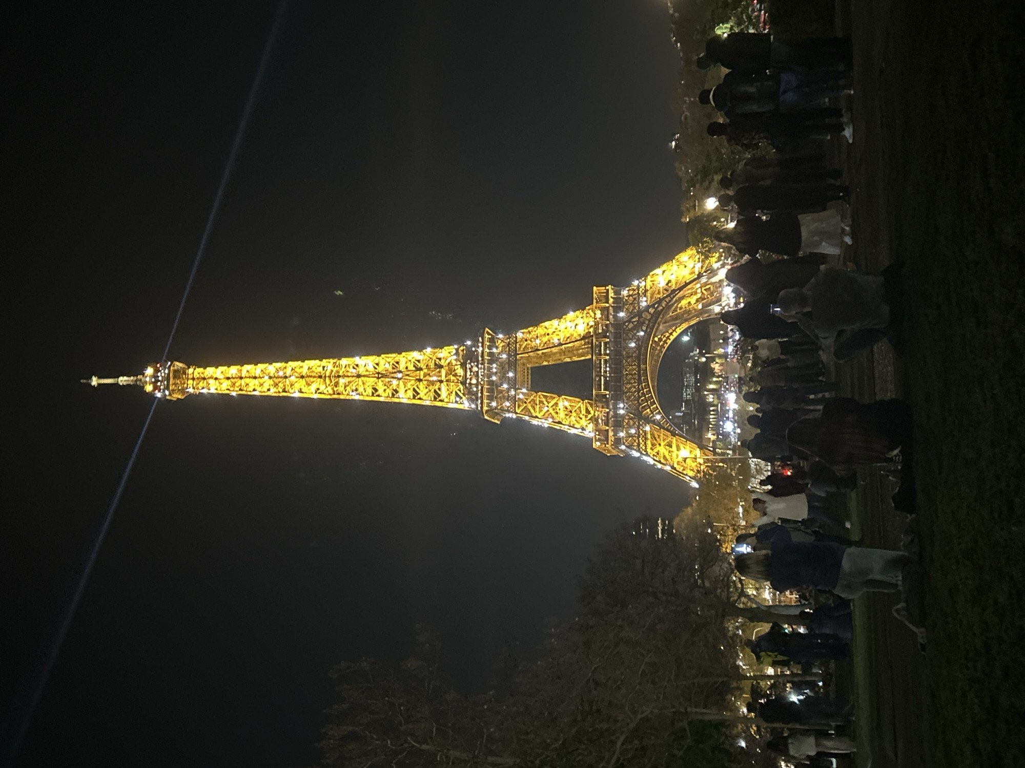 At night, the Eiffel Tower is illuminated with bright golden lights. A bright laser beam shoots up from the top of the tower into the dark sky. In the foreground are people gathered on a grassy lawn, facing the tower. Some people are taking pictures with their phones. Trees are visible in the lower left. The location is Paris, France.
