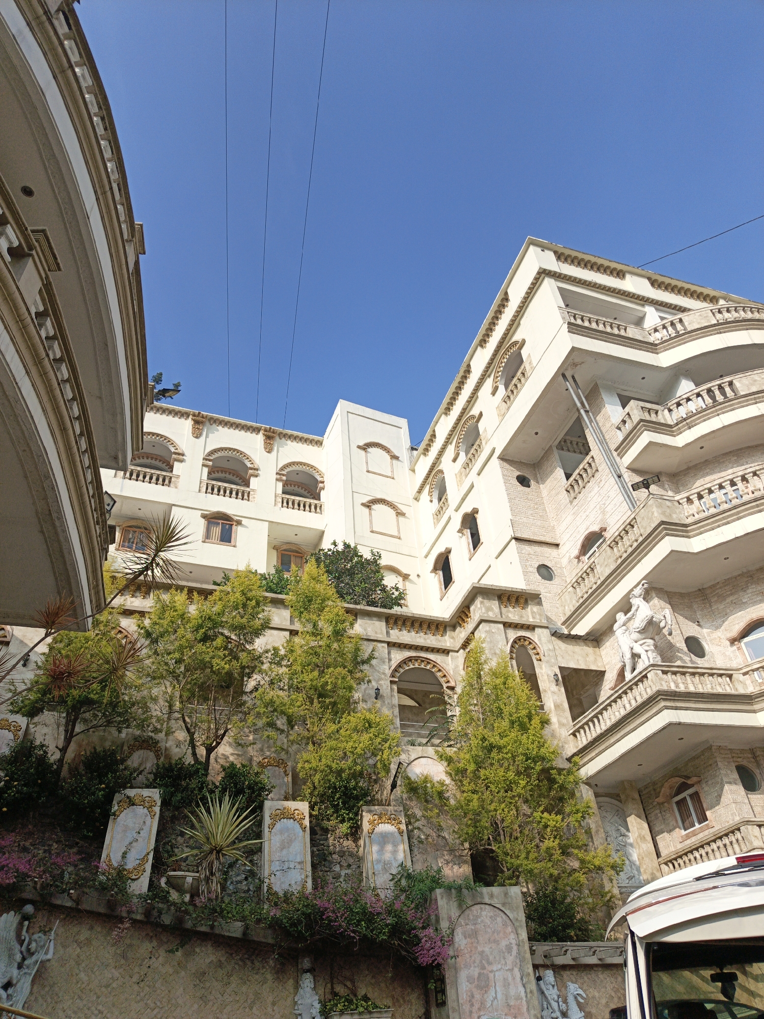 The image depicts a building with a light-colored facade, featuring multiple balconies, arched windows, and decorative elements. The building appears to be several stories high, with a complex architectural design. Lush greenery, including trees and flowering plants, surrounds the lower levels of the structure, with some plants growing on a terraced wall. Several stone-like plaques with ornate borders are visible on the wall. A white vehicle is partially visible in the lower right corner of the frame. The sky above is clear and blue. The style of the building suggests it could be a hotel, resort, or a large residential complex. While the specific location is stated to be Cibogo, Indonesia, no explicit visual cues within the image confirm this geographically. The overall impression is one of an elaborate and somewhat ornate building set within a natural, green environment.