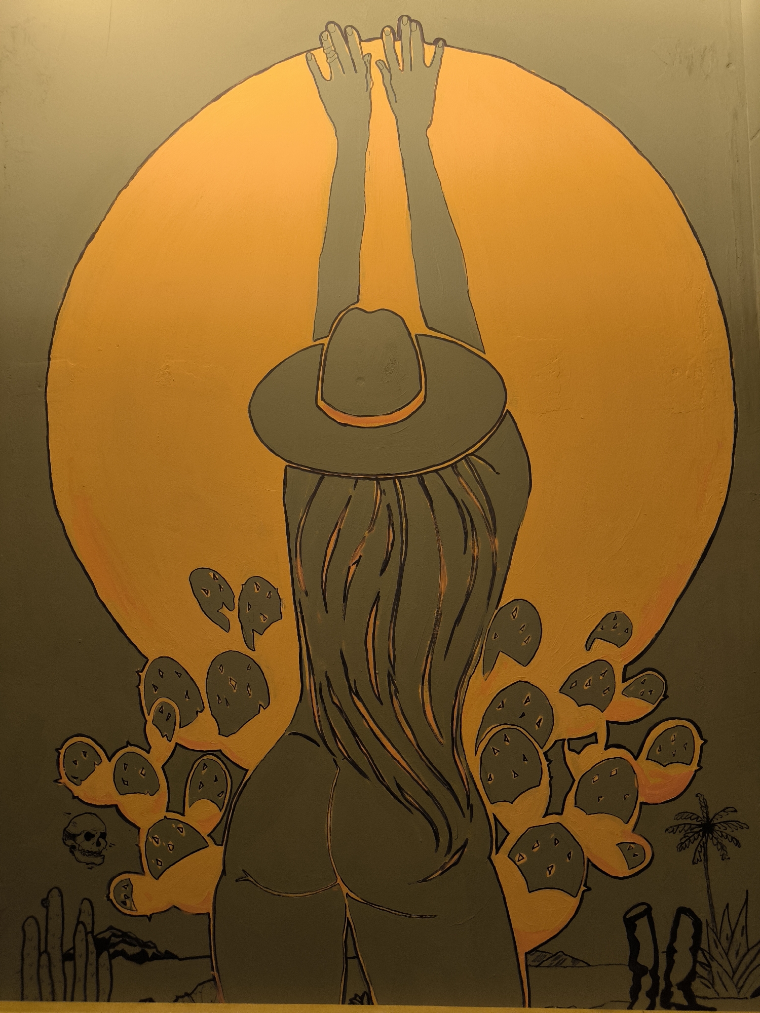 The image is a photograph of a mural painted on a wall. The mural depicts a woman wearing a cowboy hat and a long-sleeved top, viewed from behind. Her arms are raised above her head, and her long hair flows down her back. She is positioned in front of a large, orange, circular shape, possibly representing the sun or moon. To her sides and in the foreground, there are depictions of cacti and other desert flora. In the lower left corner, there is a small skull. In the lower right corner, there is a silhouette of a palm tree and other desert plants. The mural has a distinctive artistic style with bold outlines and a limited color palette of grays, oranges, and yellow.
The location of this mural in Rostov-on-Don, Russia is not provided in the image or the associated metadata. Therefore, it is impossible to determine its specific location within the city.