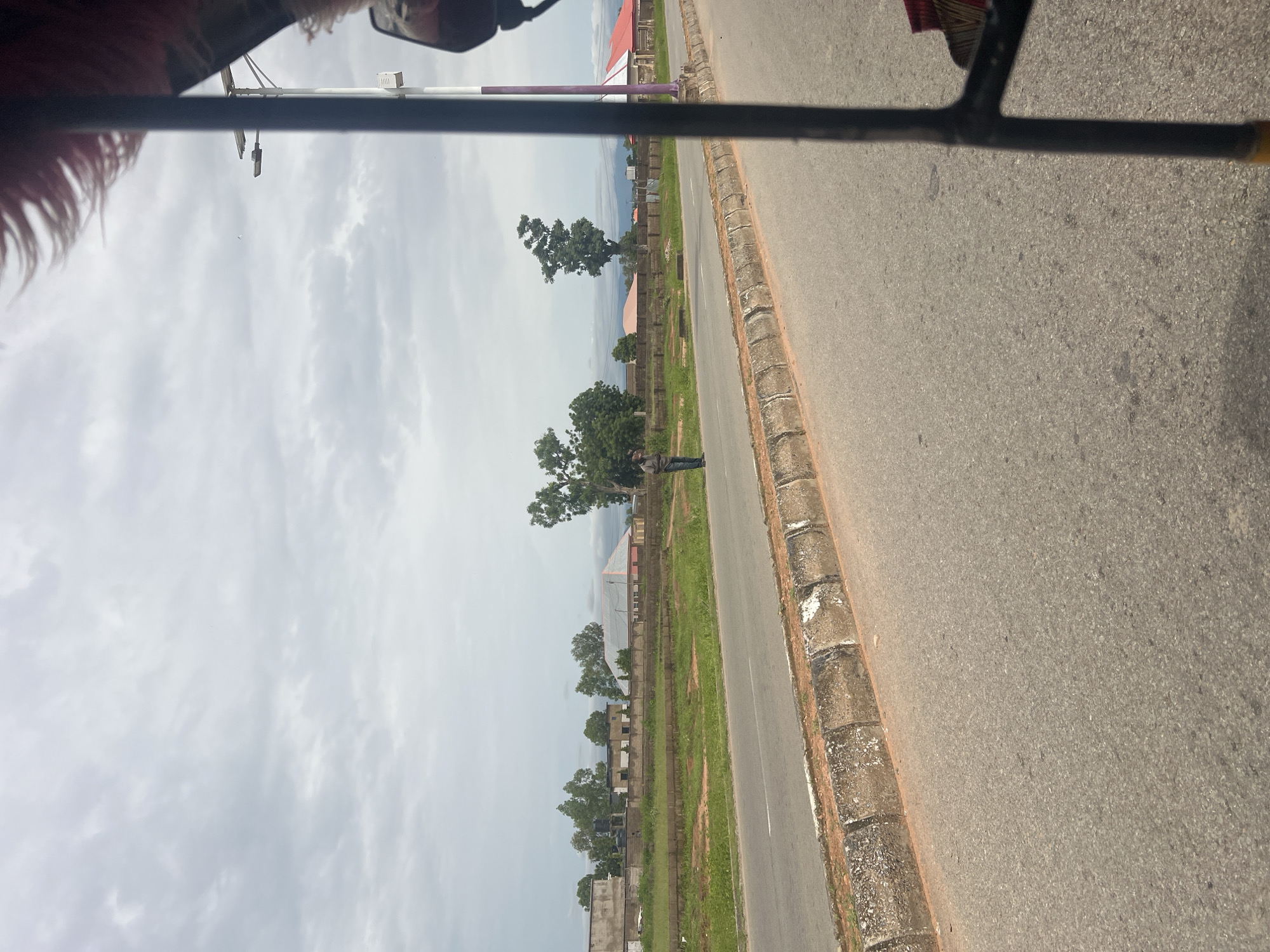 A wide shot shows a man standing on the side of a road in Kona Garu, Nigeria, with a cloudy sky overhead. The man, wearing a dark shirt and blue jeans, stands near a concrete curb. In the background, there are several buildings with red and gray roofs, surrounded by green grass and trees. A light pole with a solar panel is visible on the right side of the image. The foreground is dominated by the asphalt road and a portion of a vehicle, likely a tricycle, suggesting the photo was taken from inside it. The overall scene depicts a rural or semi-urban environment on a cloudy day.