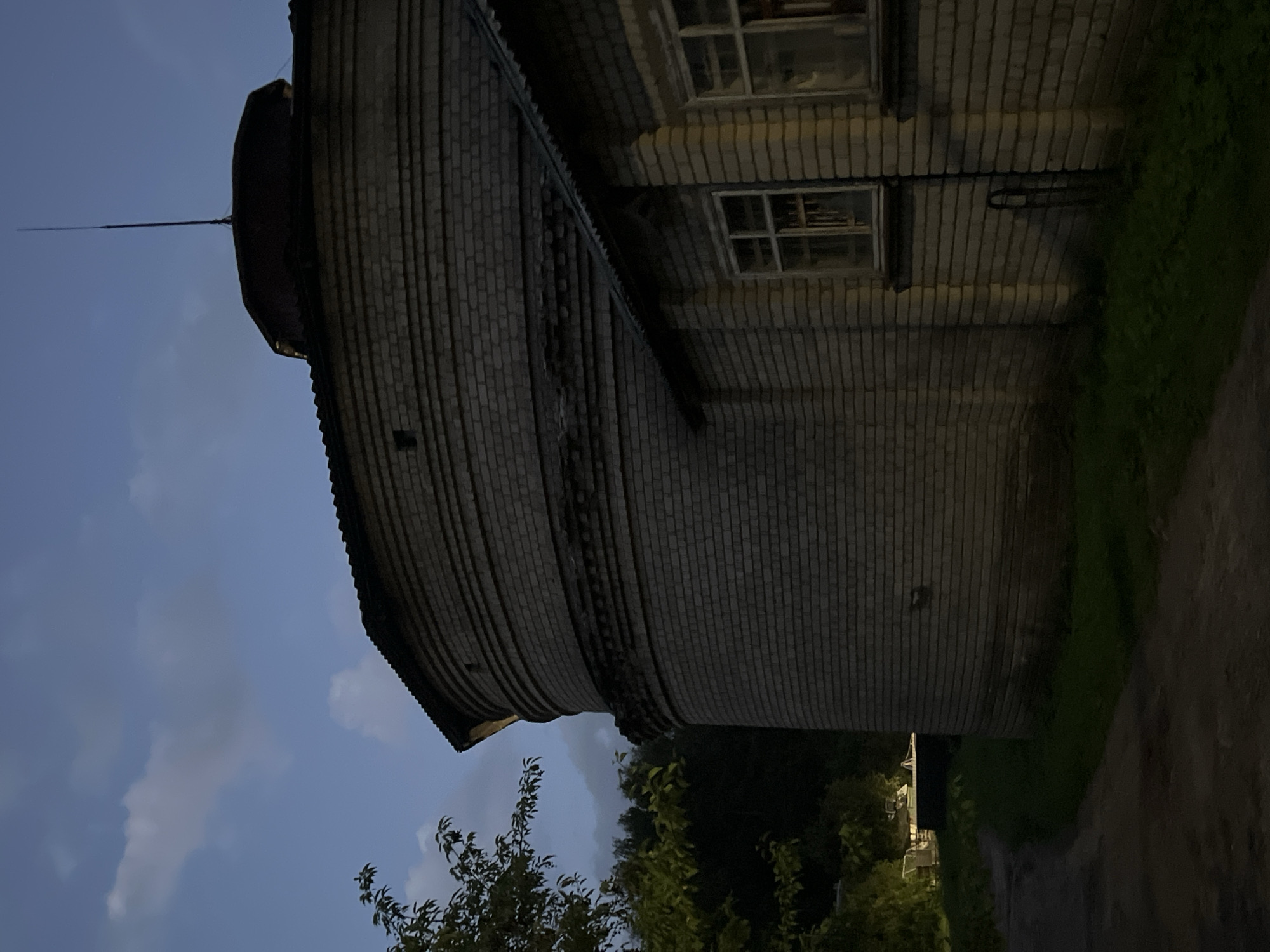 The image depicts the exterior of a circular brick building, likely a historical structure, at dusk. The building's facade is constructed from light-colored bricks laid in horizontal courses, with decorative recessed bands at intervals. A portion of a smaller, attached brick building with windows is visible to the right, connected to the main circular structure. The roof of the attached building is made of corrugated metal. A lightning rod extends from the top of the circular building. The surrounding environment includes a dirt path leading away from the building, grass, and trees, with a dark, clouded sky overhead. The scene is dimly lit, suggesting it is either early morning or evening. There are no people or visible actions taking place. The context provided in the prompt about the location being Vilnius, Lithuania, is not visually verifiable from the image itself.