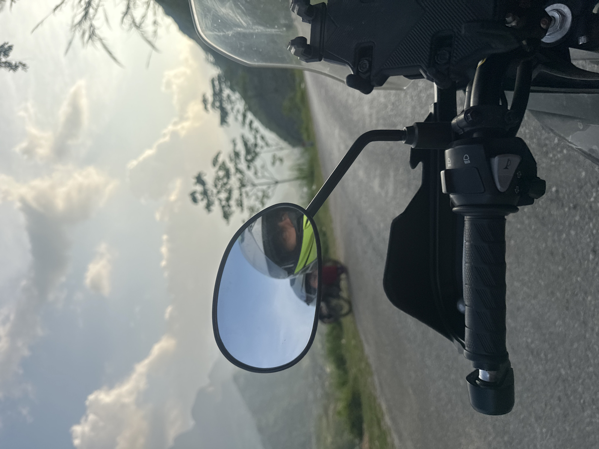 A close-up shot shows the handlebars of a motorcycle, including the right-side grip and mirror. The mirror reflects the image of the road behind and a helmeted rider. The scene is set in Du Già Commune, Vietnam, as implied by the background landscape of mountains and foliage. The motorcycle is positioned on a road surface.