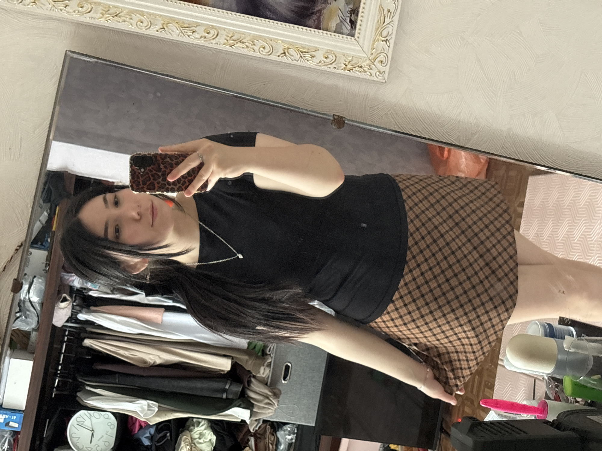 A female is taking a selfie in a mirror. The mirror is hanging on a wall with patterned wallpaper, below a framed painting. She is holding a phone with a leopard-print case. She is wearing a black shirt, a short plaid skirt, and a silver necklace. Her dark hair is pulled to one side. A closet filled with clothes, and a clock reading approximately 1:10, is visible in the mirror's reflection, along with various objects on a table. There is no indication of the location being Izhevsk, Russia.