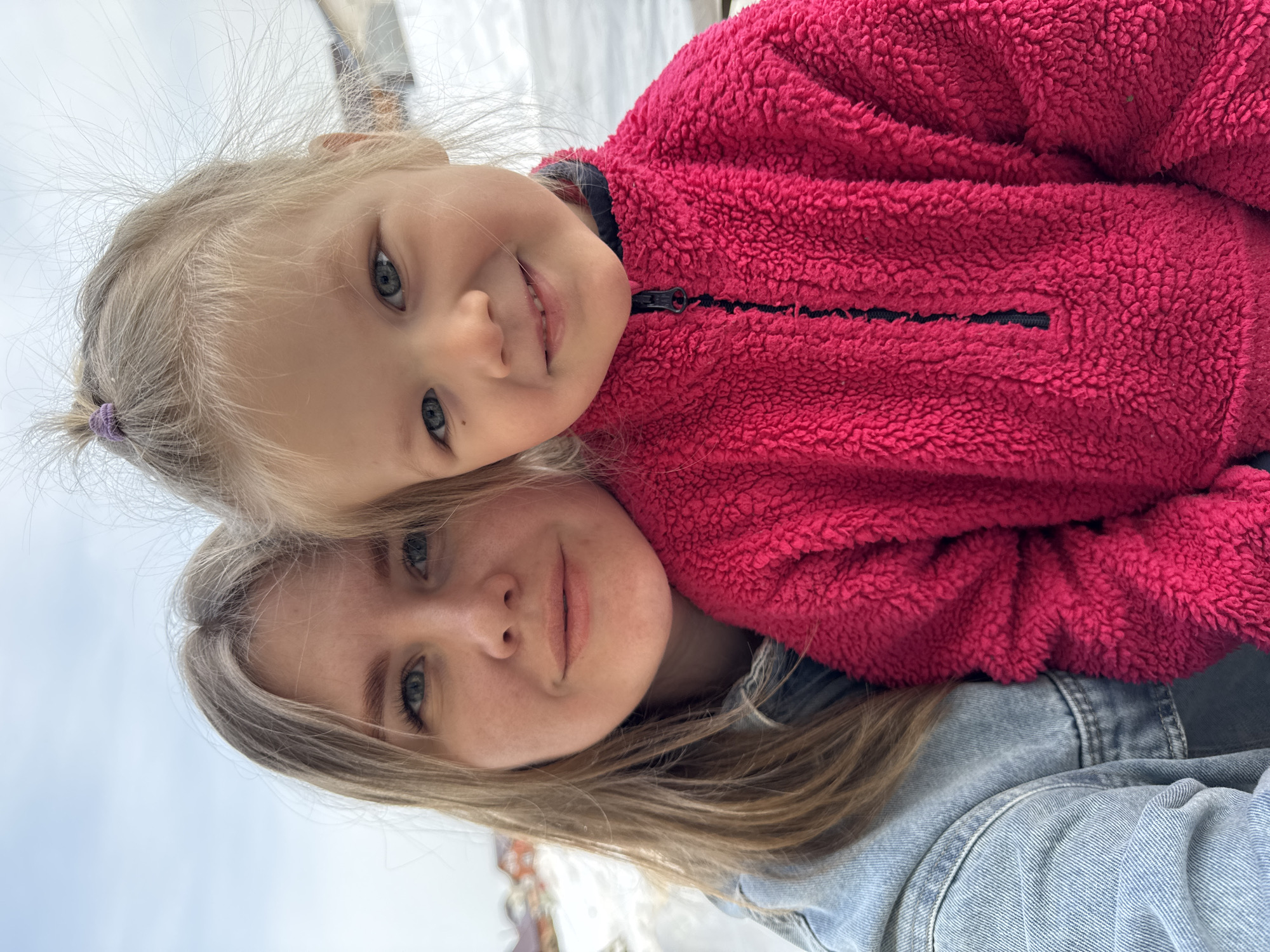A close-up, selfie-style portrait captured in Gornyi (Dvurechenskoe S/P), Russia, features an adult female and a young female child. The adult female, with long blonde hair and blue eyes, is positioned on the left, wearing a light blue denim jacket. The child, approximately 3-5 years old, with blonde hair tied in a small top ponytail secured by a purple elastic, blue eyes, and visible developing or missing front teeth, is positioned to the right and slightly in front of the adult. The child is wearing a textured bright fuchsia fleece jacket with a black quarter-zip and dark collar. Both subjects are looking directly at the camera with slight smiles. The background is blurred, revealing patches of snow on the ground and indistinct residential structures with orange roofs in the distance under a light blue, overcast sky.
