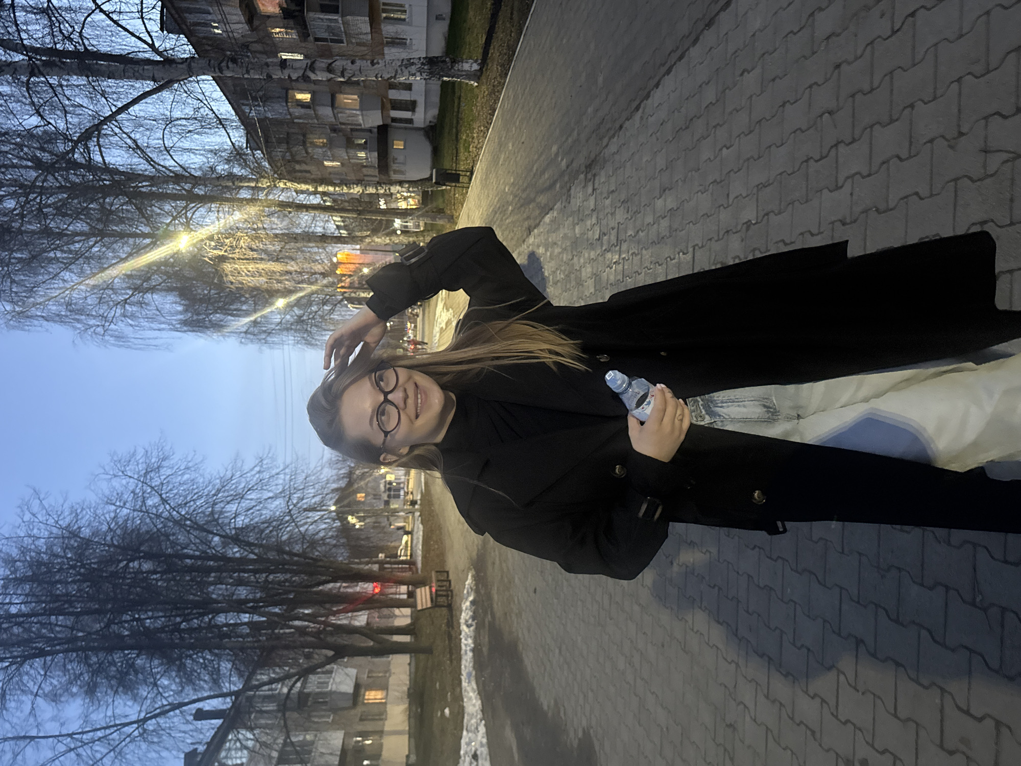 A woman stands on a brick-paved sidewalk in Chaikovsky, Russia. She is wearing glasses and a dark coat. She is holding a water bottle in her left hand and has her right hand up near her hair. The setting appears to be outdoors with buildings and bare trees visible in the background, and it looks to be dusk or early evening.