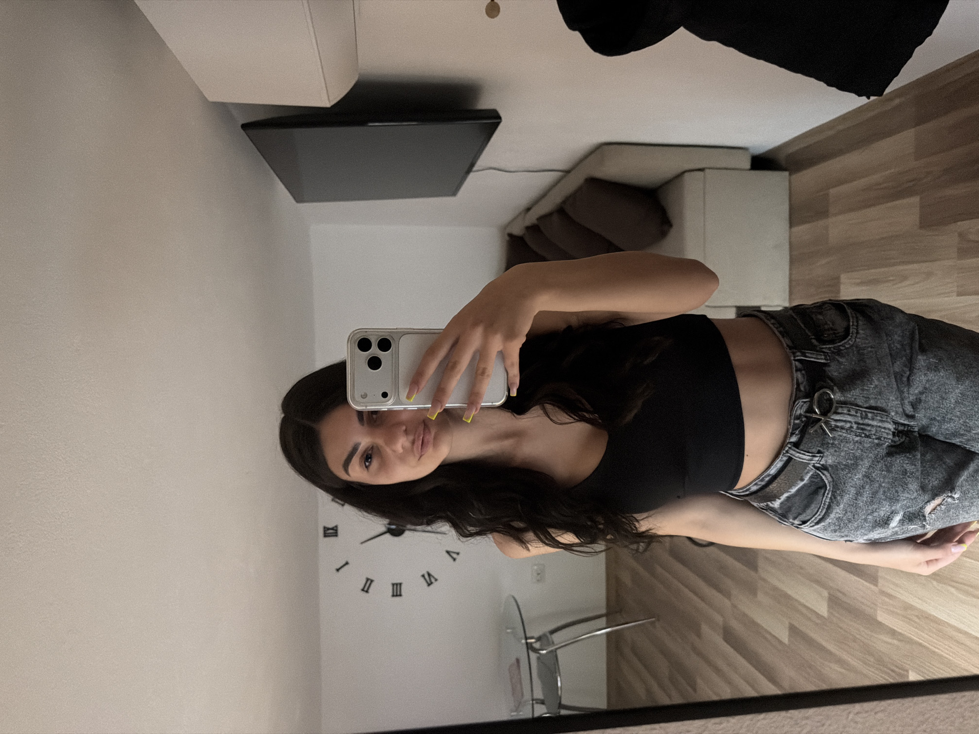 A young woman is captured in a mirror selfie, standing in what appears to be a modern living space in Gabrovo, Bulgaria. She has long, dark, wavy hair and is looking directly towards the camera with a subtle smile. She is wearing a black crop top and high-waisted, acid-wash grey jeans that feature a silver ring on the front belt loop area. Her fingernails are painted a vibrant yellow. She holds a light-colored smartphone with a clear case in her right hand to take the photo.
The background of the room includes light-colored walls and a wooden floor with a diagonal pattern. On the wall to her left, a large, decorative wall clock with black Roman numerals (I, II, IV, V, VII, VIII, X, XI clearly visible) is mounted. Partially visible behind her is a clear glass-top table with metal legs and a matching chair. Further into the room, a wall-mounted flat-screen television or monitor is seen, along with a white sofa adorned with dark cushions and a simple white console or cabinet. The room is well-lit, presenting a contemporary and casually furnished interior.