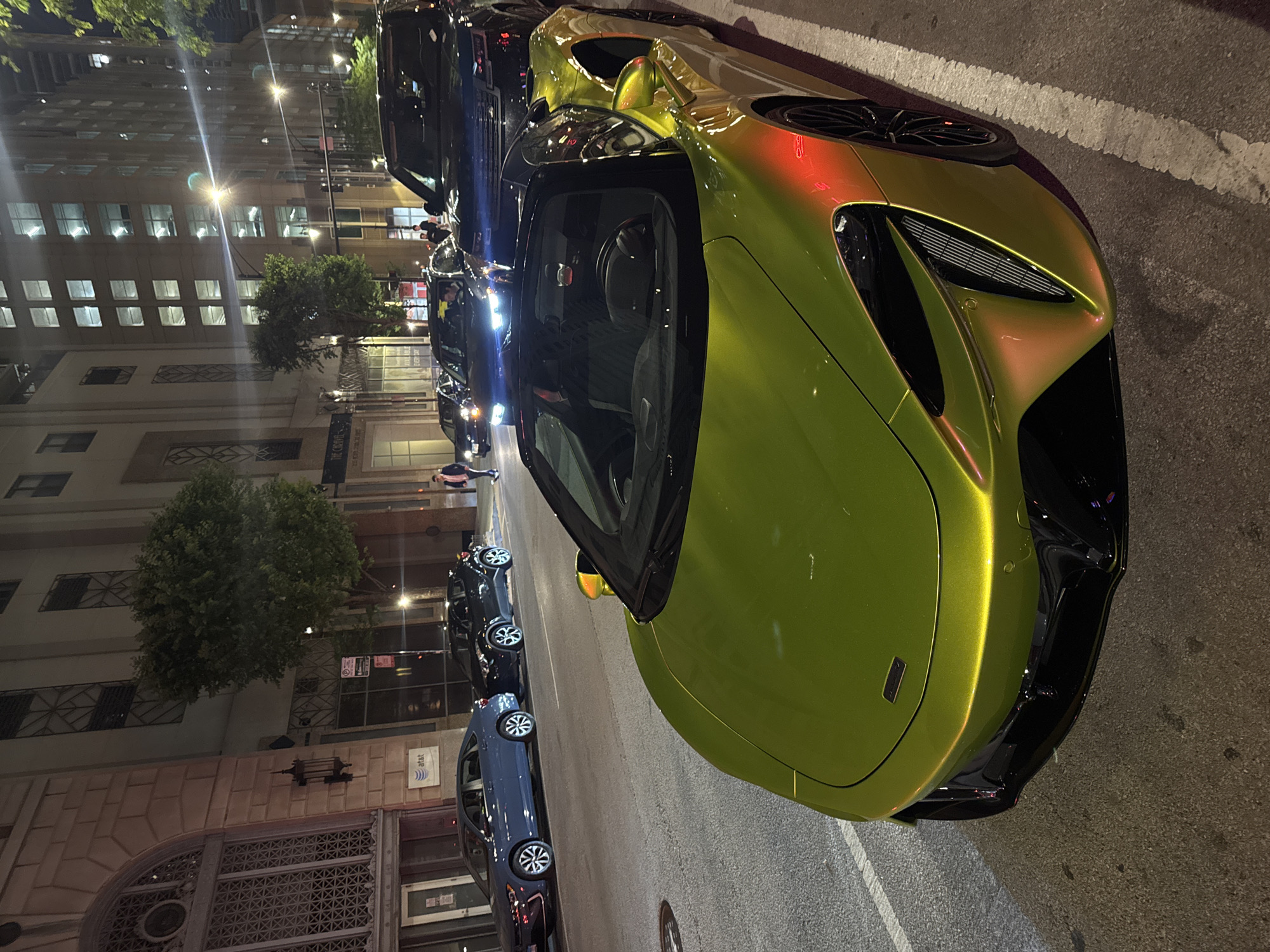 A nighttime street scene in Chicago, Illinois features a row of parked cars alongside a building with illuminated windows. A green and gold McLaren sports car dominates the foreground. Other cars, including a blue sedan and black SUVs, are parked behind the McLaren. The building on the left has architectural details and a sign partially visible. Streetlights illuminate the scene. Pedestrians can be seen in the distance walking along the sidewalk.