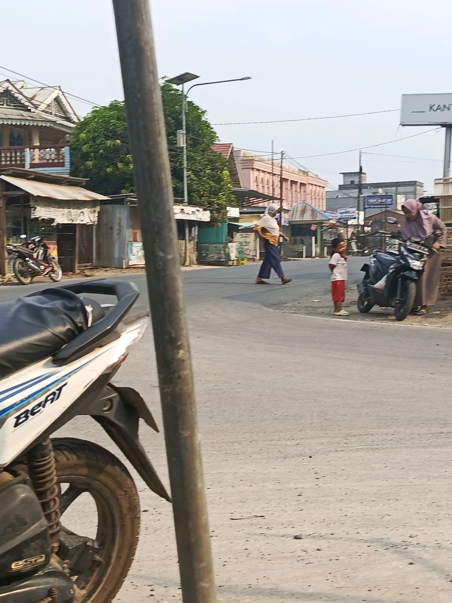 In Megang Sakti I, Indonesia, a street scene unfolds with a light pole in the foreground, partially obscuring a white scooter with blue accents. On the street, a person wearing a white head covering and a yellow garment walks to the left, their body angled away from the camera. To their right, a young child in a white t-shirt and red shorts stands facing the scooter. A woman in a pink headscarf stands next to a black scooter, appearing to interact with the child. In the background, buildings with pitched roofs and intricate wooden trim are visible, along with other structures, trees, and overhead wires. A sign reading "_ KANTOR" is mounted on a pole in the distance. The overall environment suggests a public area, possibly a market or residential street, with signs of daily life and commerce.