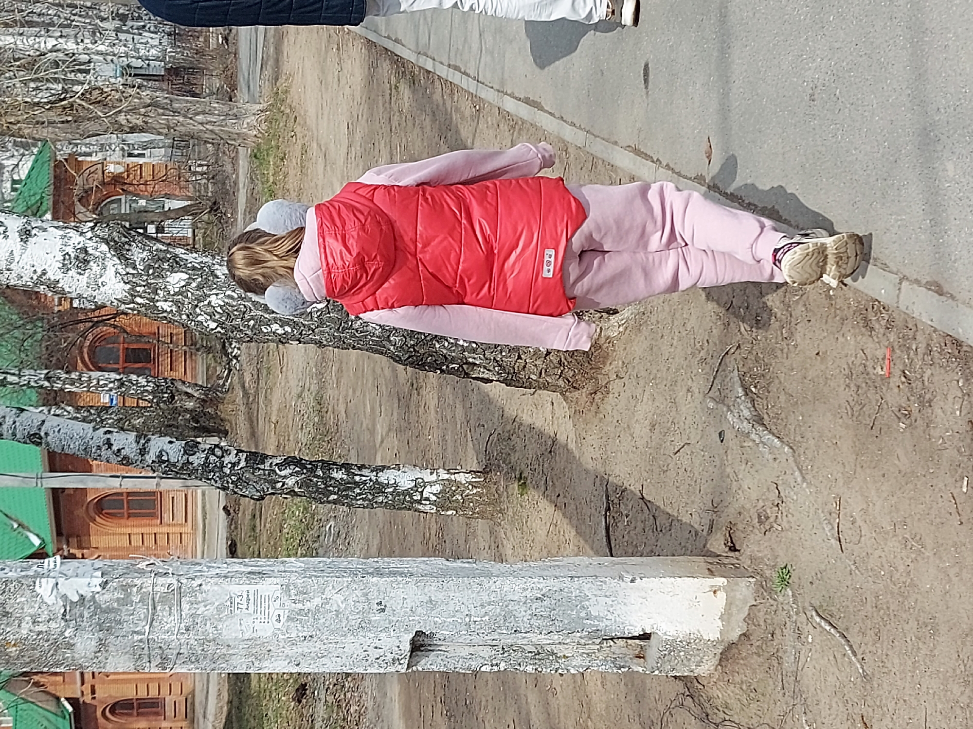 The image depicts an individual wearing a pink sweatsuit and a red vest, standing on a sidewalk next to a dirt area with birch trees and a concrete pole. Buildings with brick facades and green roofs are visible in the background. The photograph was taken in Chaikovsky, Russia.
