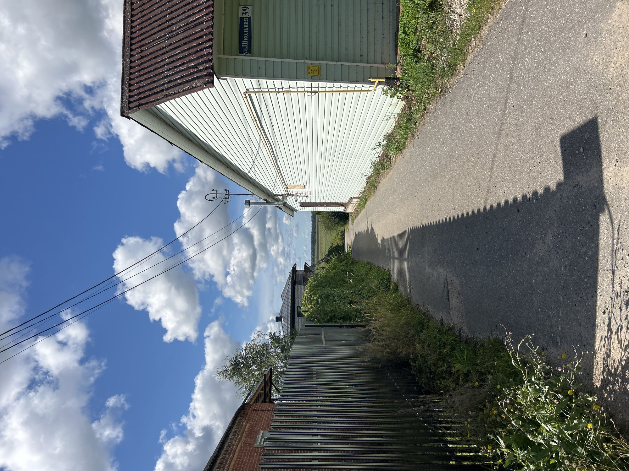 The image depicts a narrow asphalt pathway leading through a rural area, possibly within the city of Kalitsino, Russia, given the presence of a sign reading "ул. Школьная 39" (Shkolnaya Street 39). On the right side of the path is a light green, horizontally paneled building with a dark brown, corrugated roof. A yellow gas pipe runs vertically along the side of this building, connecting to an overhead line. A blue street sign with white lettering is affixed to the corner of the building. On the left side of the path, a tall, dark metal fence runs alongside, with a red brick building visible behind it and a mature tree with sparse foliage. Beyond the fence and to the left, dense green foliage and wild grasses grow along the path's edge. The path is partially shaded by the fence, casting a distinct serrated shadow. In the distance, a flat, open field is visible under a bright blue sky dotted with fluffy white clouds. No people are present in the scene. The environment appears to be a sunny day with moderate cloud cover.