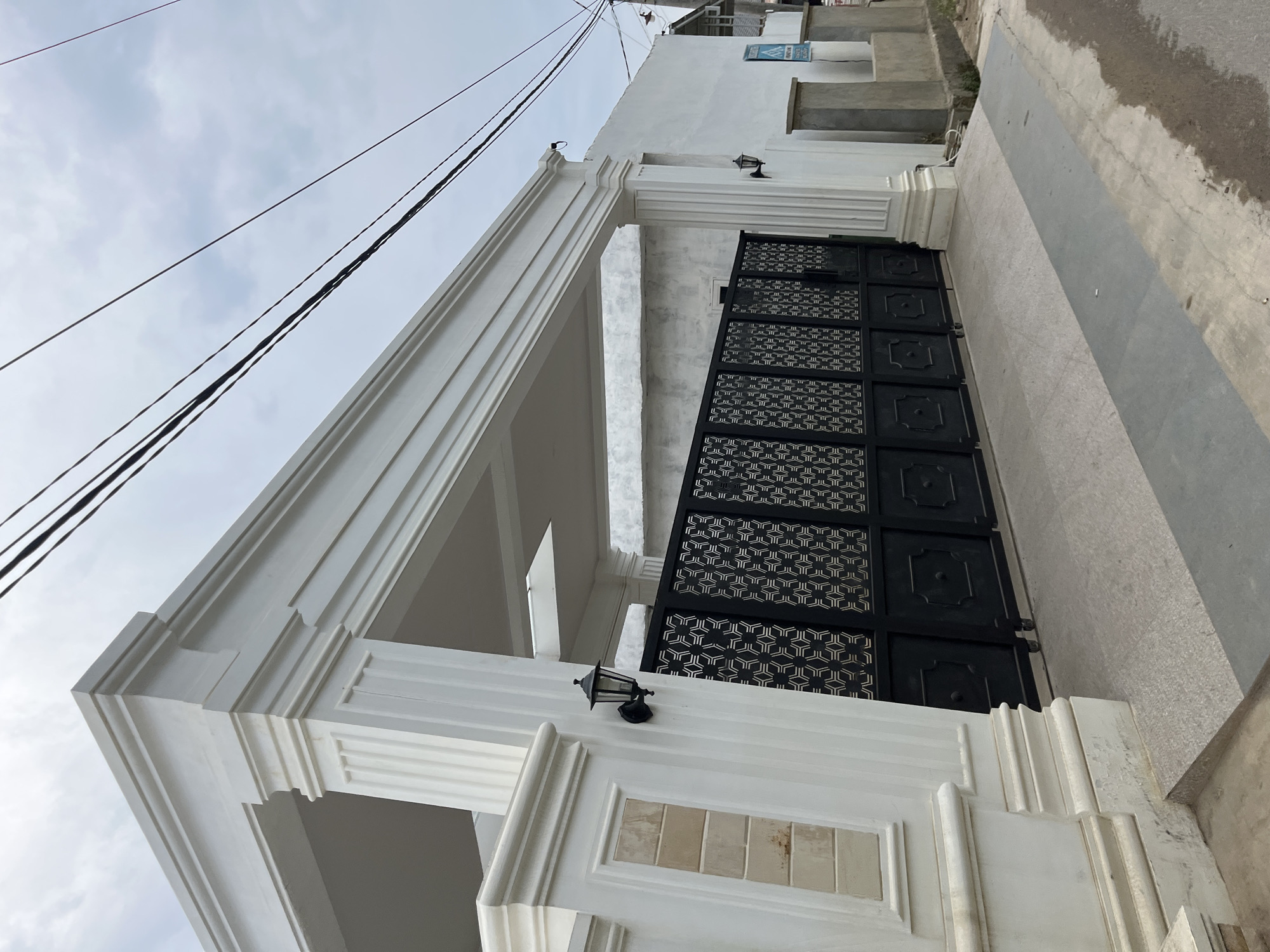 The image depicts a modern white gate and facade in Tanahterban, Indonesia. The gate is black with an intricate geometric pattern and is flanked by large white pillars. A white awning with decorative molding extends over the entrance. An outdoor lamp is mounted on the left pillar. In the background, a white wall runs alongside the property, with a street and sidewalk visible in the foreground. Overhead, black power lines are strung against a cloudy sky. The overall impression is one of a well-maintained, upscale residence.