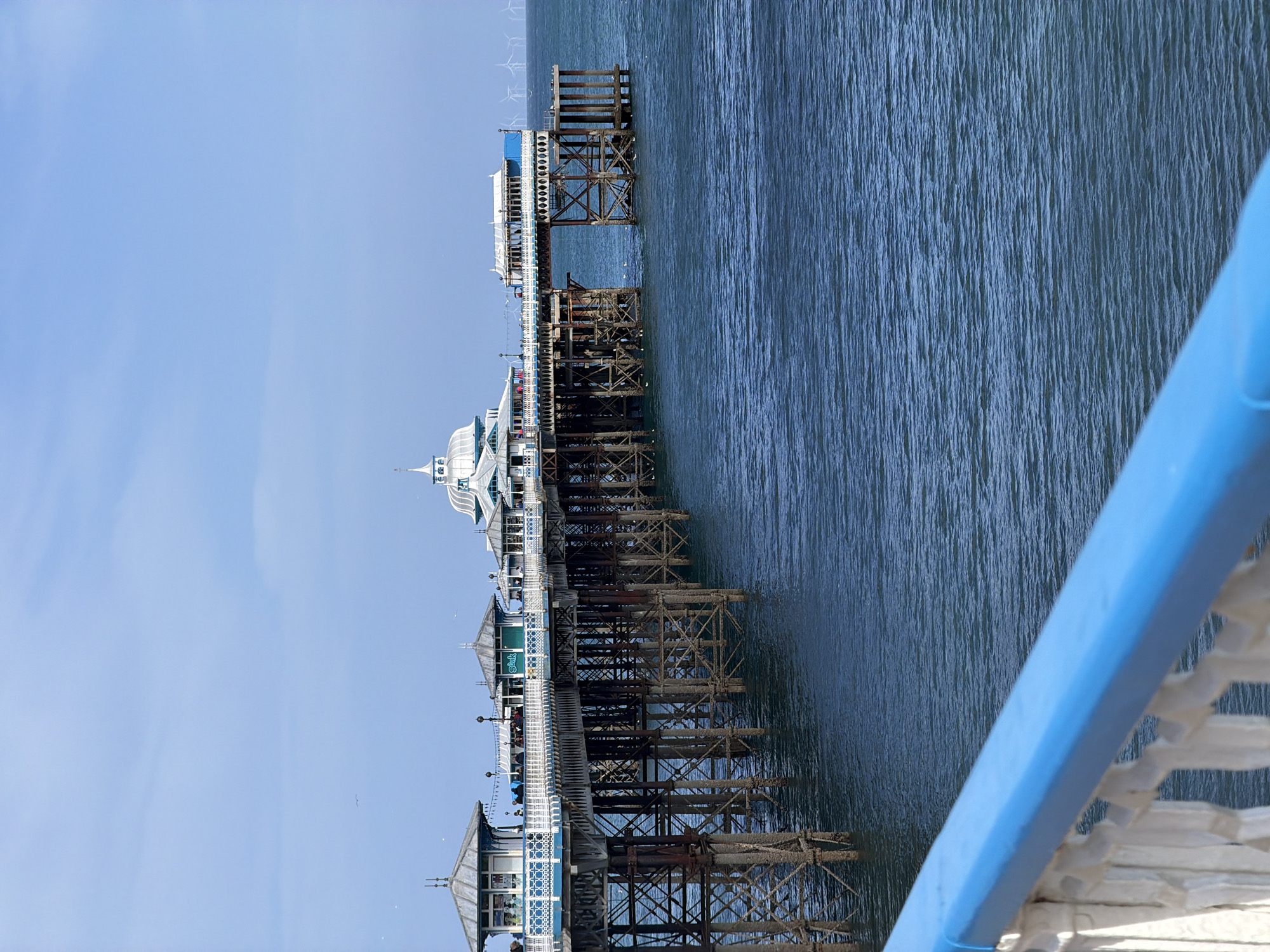The image depicts a view of Llandudno Pier in Llandudno, United Kingdom, from a slightly elevated perspective on the pier itself. The pier extends horizontally across the frame, supported by a complex network of metal and wooden stilts that are partially submerged in the sea. The water is a rippled dark blue. The pier structures feature decorative elements, including ornate pavilions and railings. A blue railing is prominent in the foreground on the right. The sky is a clear, pale blue. In the far distance at the end of the pier are turbines. There are no people visible in the frame.